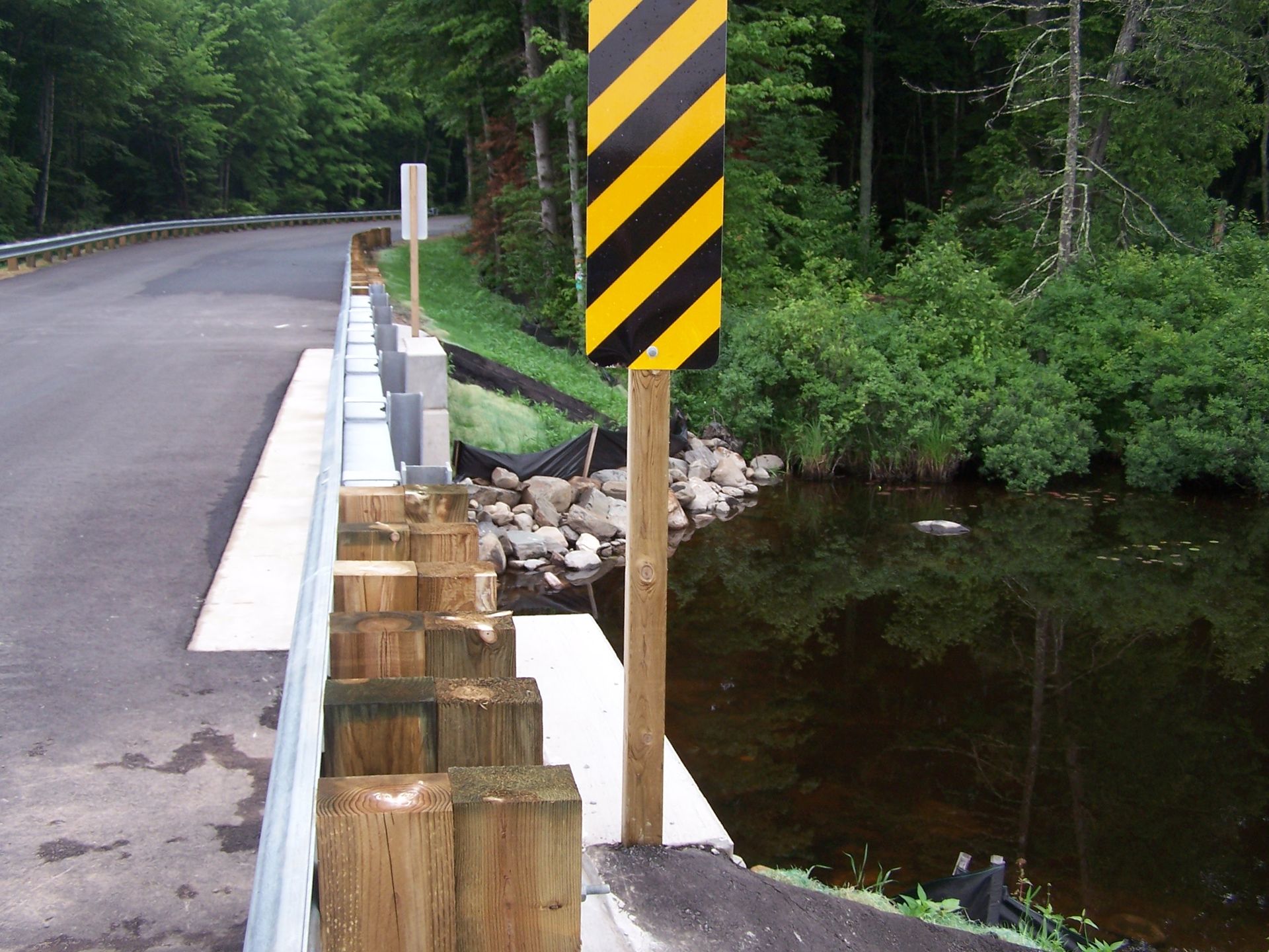 A road bridge with wooden guardrails next to a water body, featuring a yellow and black diagonal hazard warning sign.