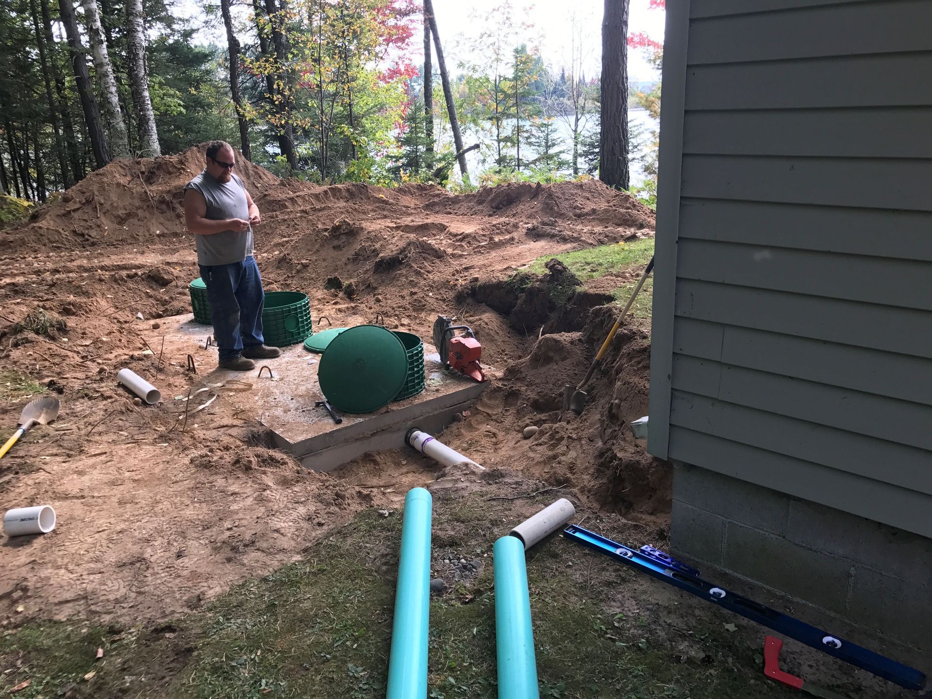A person in a work site beside a house, standing near an exposed septic tank with green lids and PVC pipe segments.