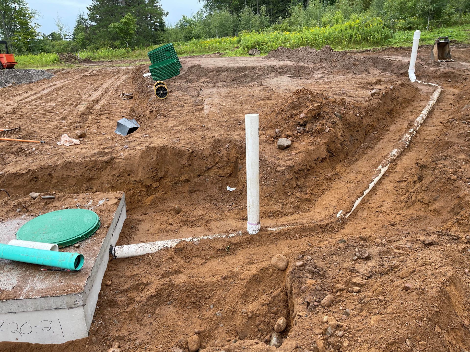 A construction site showing a concrete septic tank with a green lid, connected to white PVC pipes in trenches.