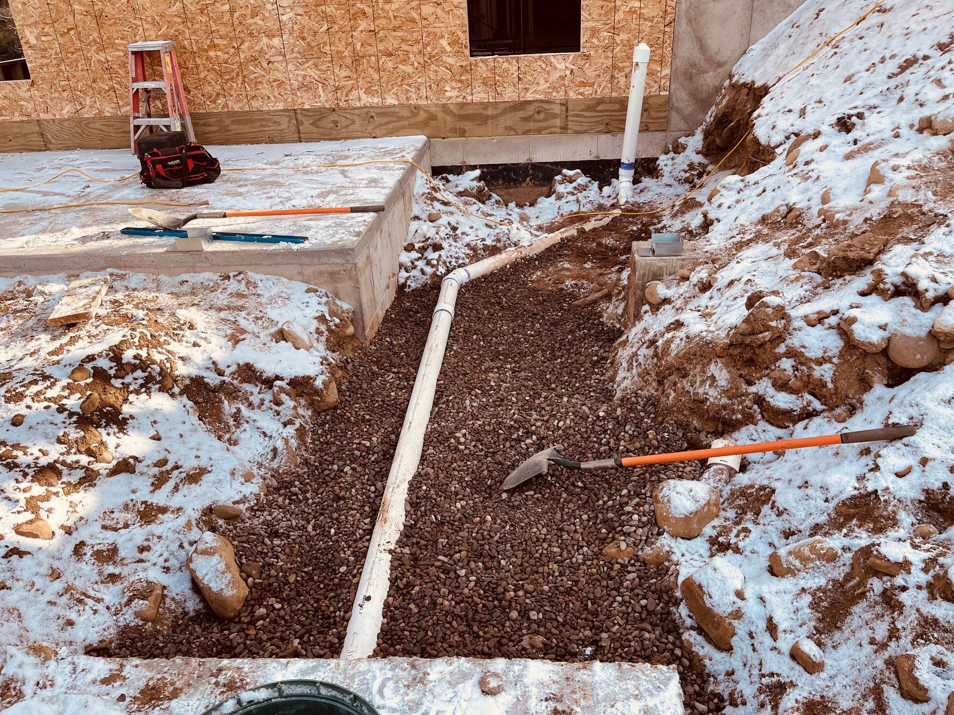 A construction site featuring a white pipe in a gravel-filled trench alongside a building foundation under light snow. A construction site featuring a white pipe in a gravel-filled trench alongside a building foundation under light snow.