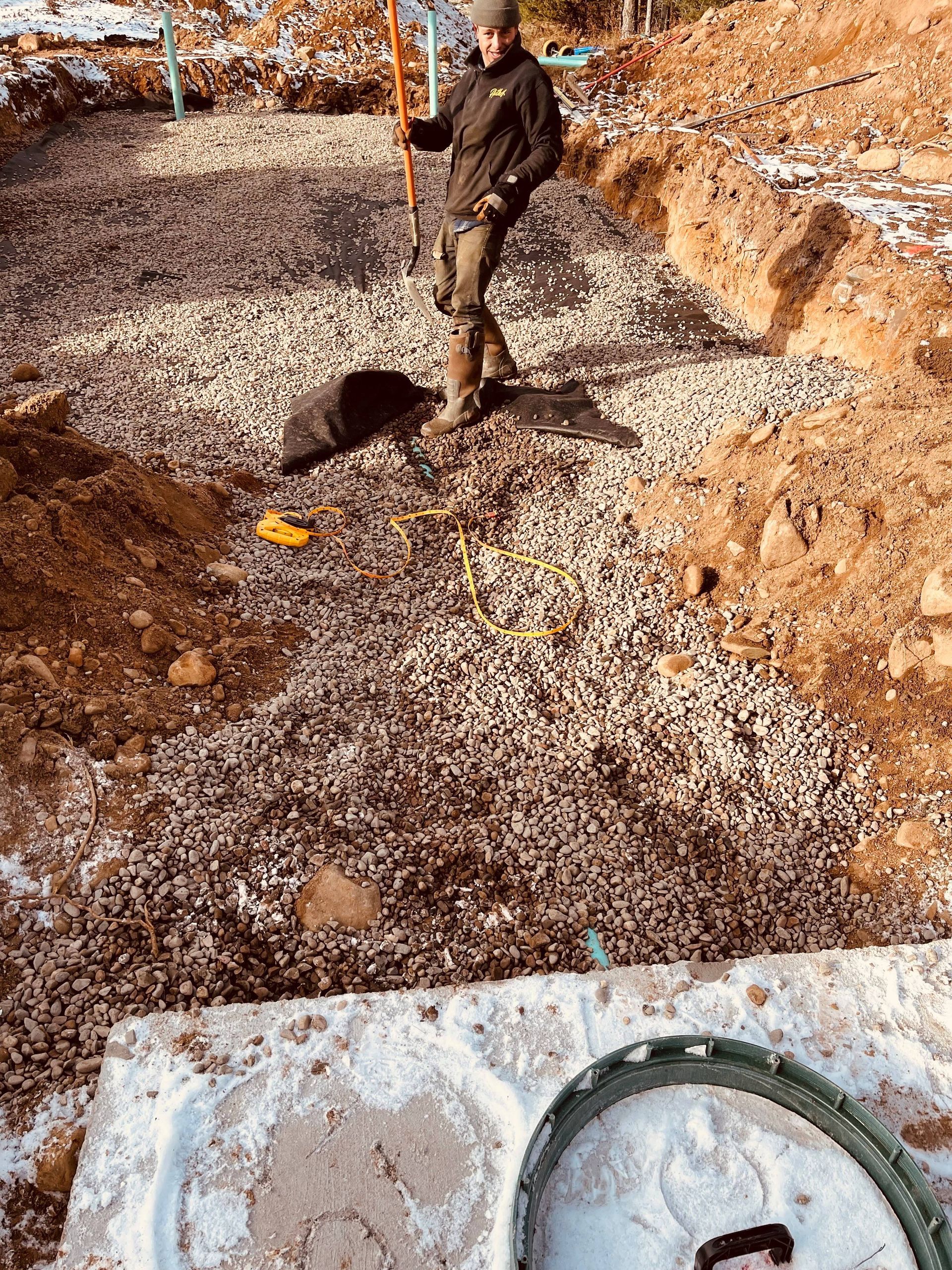 A person in work gear stands in a deep, gravel-filled excavation trench with snow on the surrounding earth.