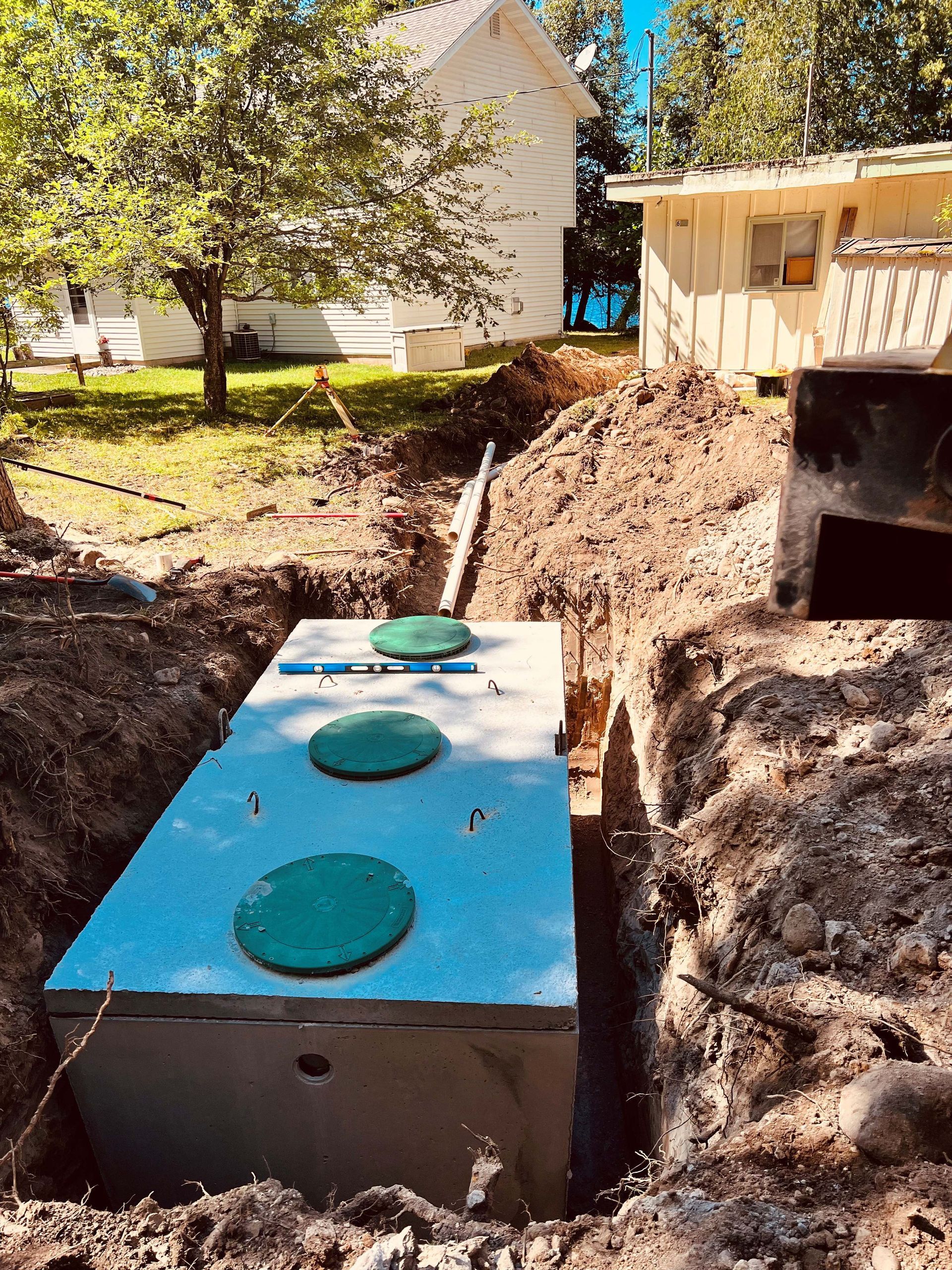 A newly installed rectangular concrete septic tank with three green circular lids sits in a dirt trench near two houses. A newly installed rectangular concrete septic tank with three green circular lids sits in a dirt trench near two houses.