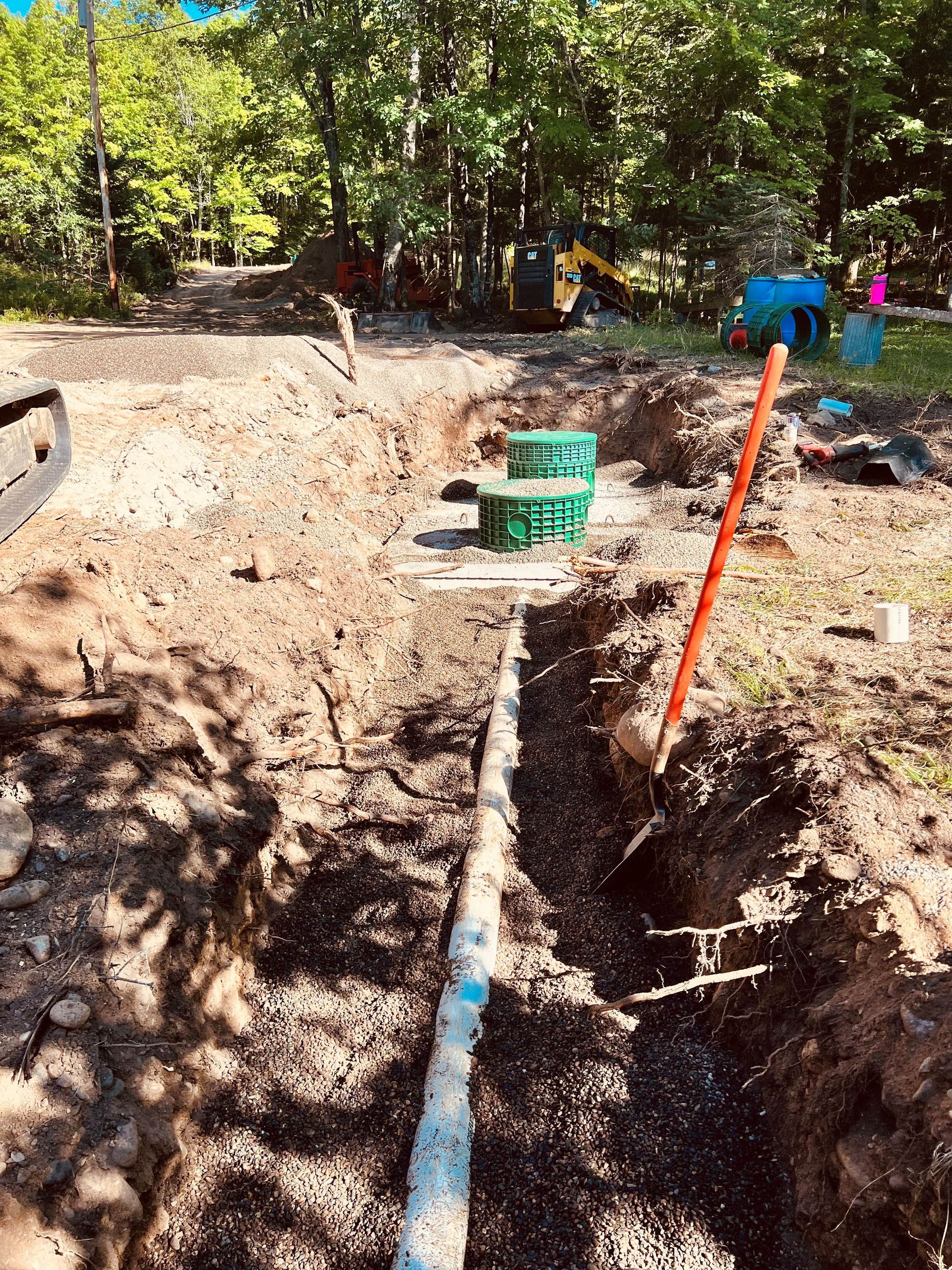 A trench with a pipe running through the center, leading to two green septic access covers in a wooded construction site.