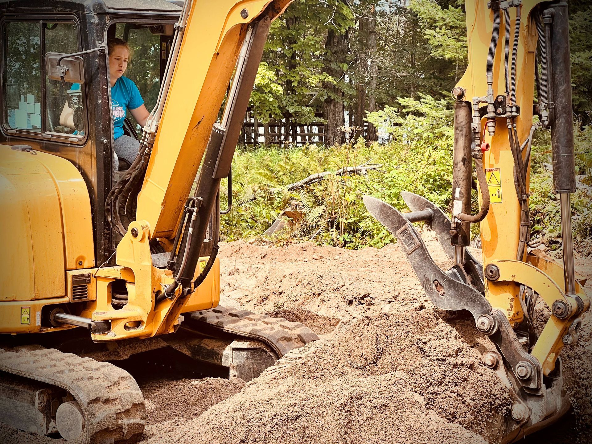 A person in a blue shirt operating a yellow excavator in a dirt-filled, wooded area. A person in a blue shirt operating a yellow excavator in a dirt-filled, wooded area.