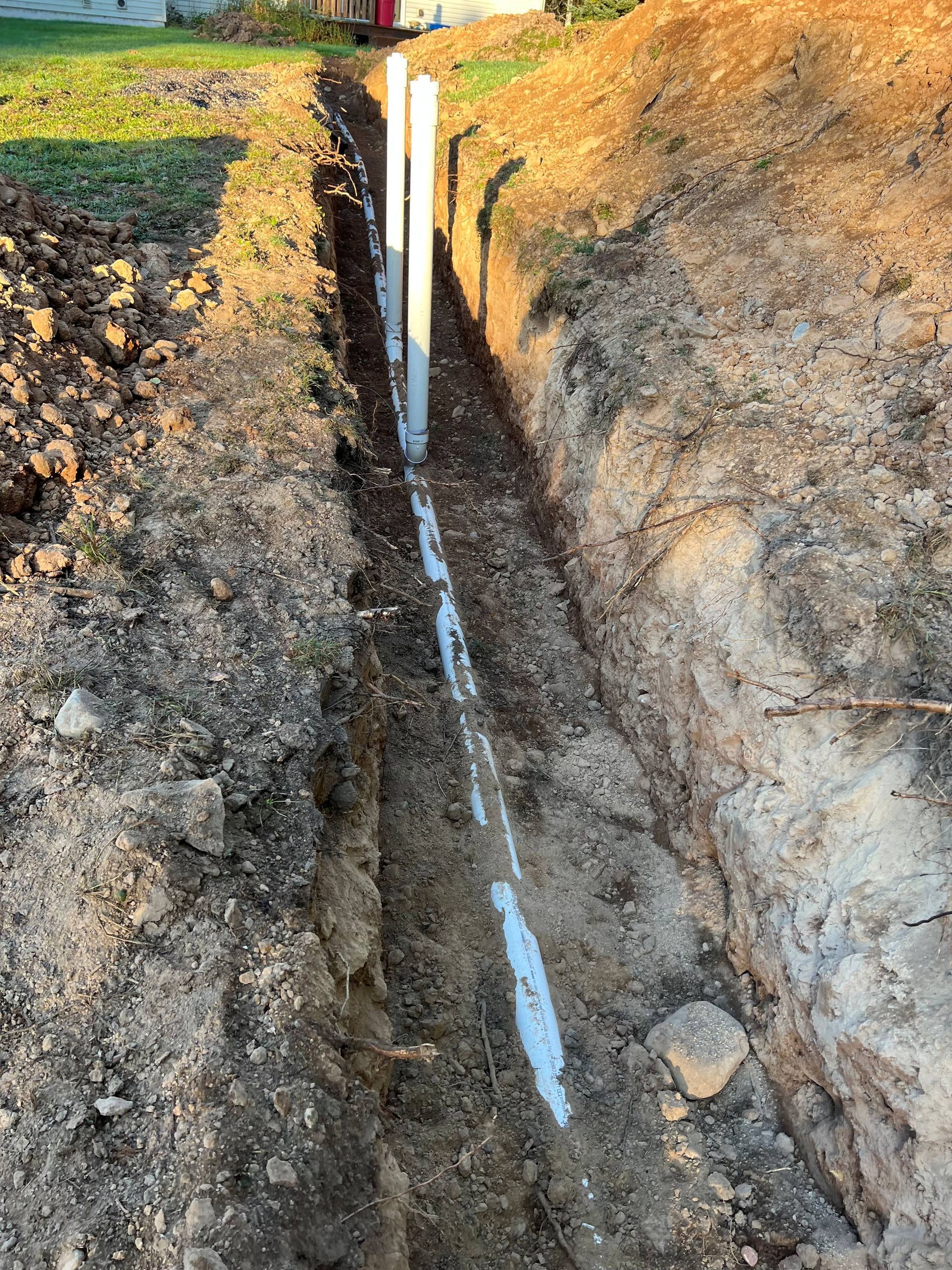 A narrow trench in the ground containing white PVC pipes extending vertically from a horizontal main pipe.
