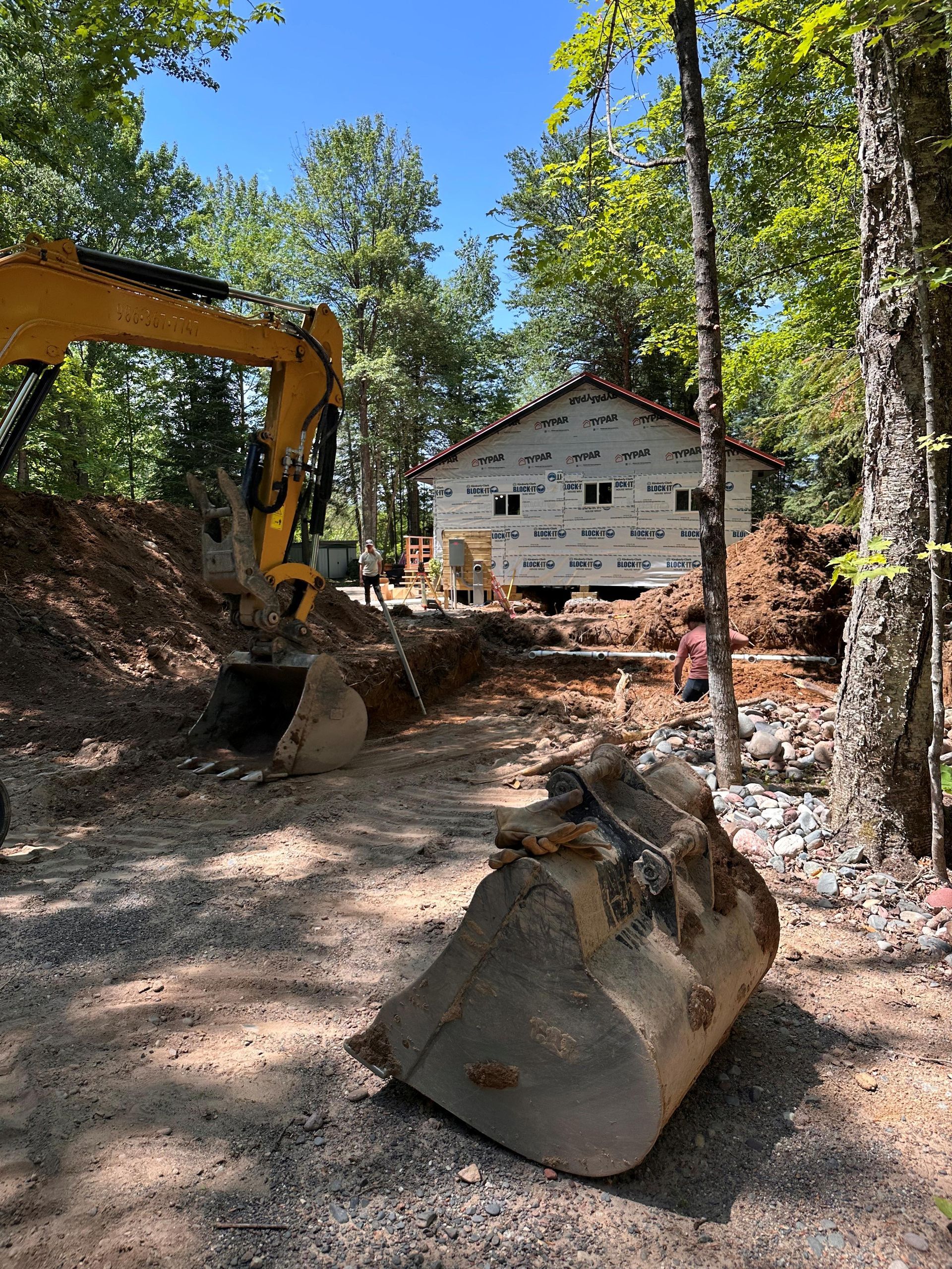 A yellow excavator works in a dirt clearing near a house under construction with white exterior sheeting.