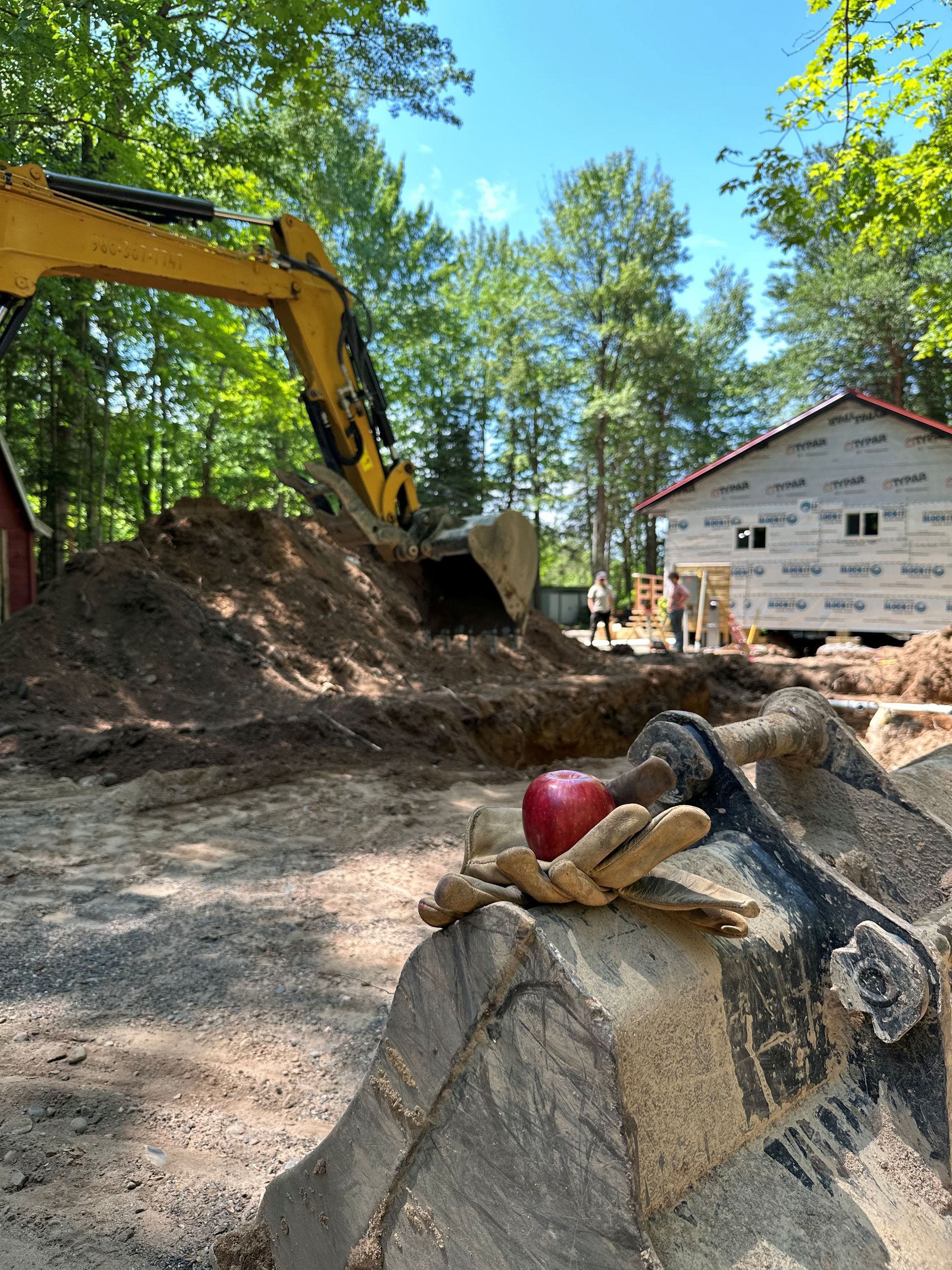 An apple rests on a tan work glove placed atop a yellow excavator bucket at a construction site with a house under way.