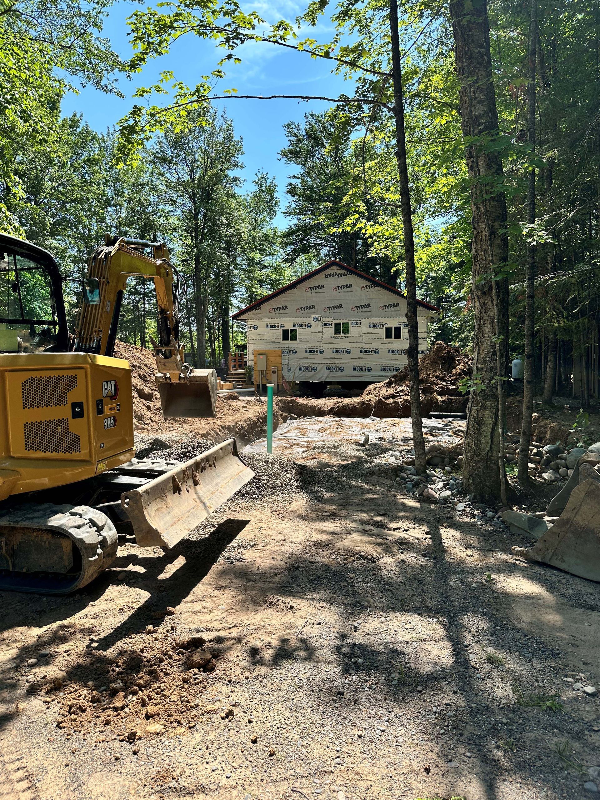 A yellow excavator sits in a clearing in front of a white cabin undergoing renovations in a wooded area. A yellow excavator sits in a clearing in front of a white cabin undergoing renovations in a wooded area.