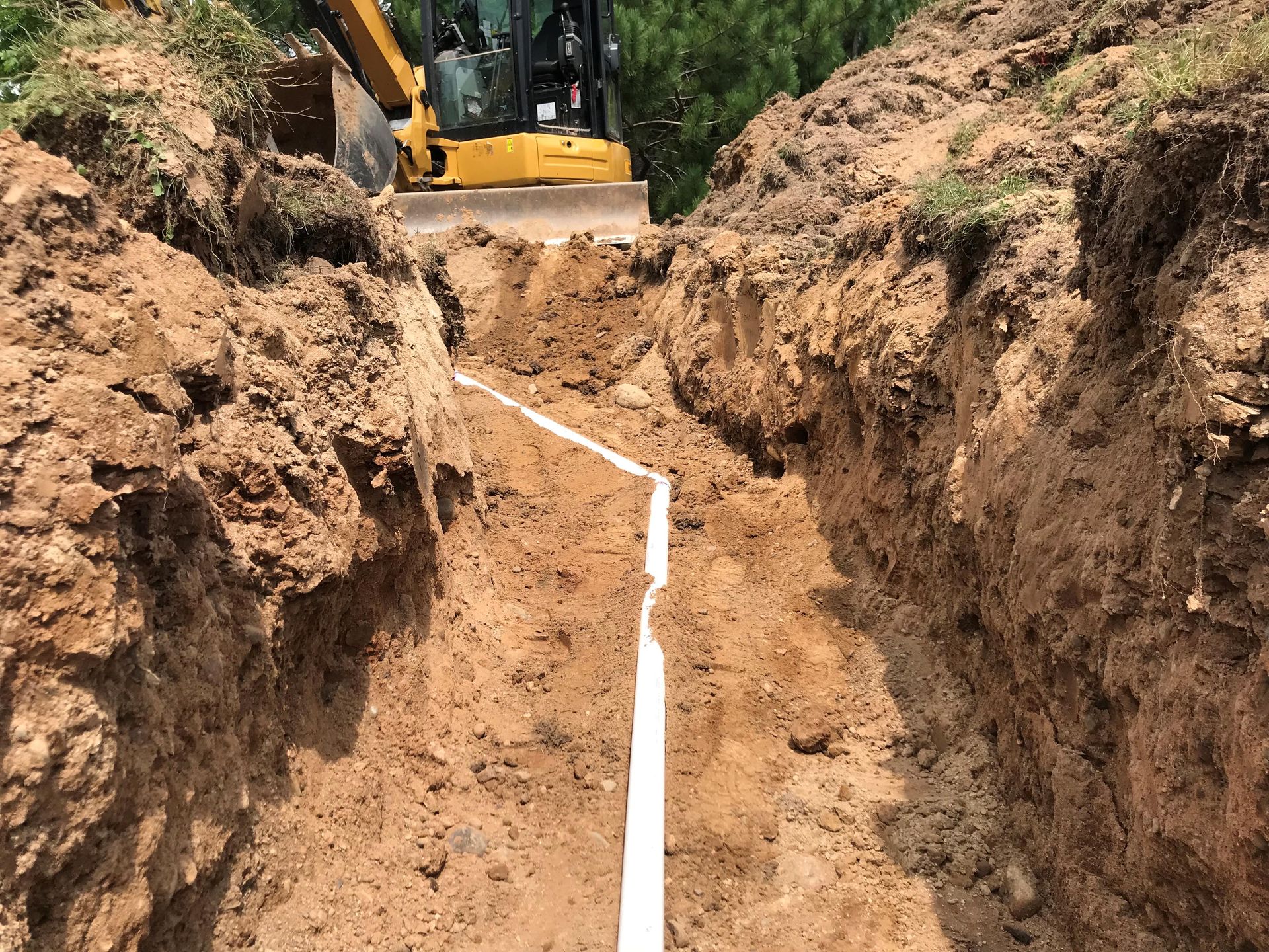 A yellow excavator parked at the edge of a deep trench containing a long, white plastic pipe in a dirt landscape.