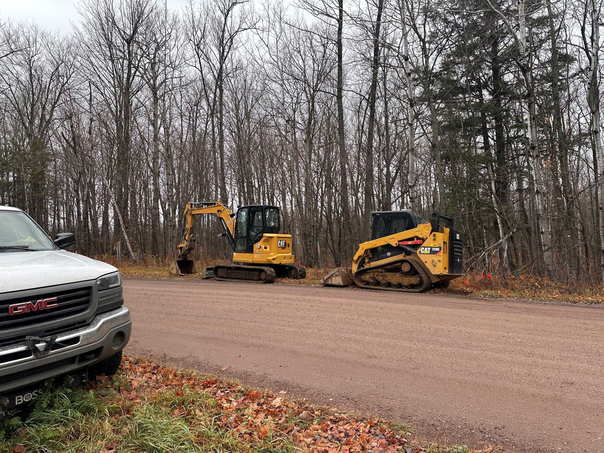 Two yellow Caterpillar construction machines parked on a gravel road next to a GMC truck in a forest setting.