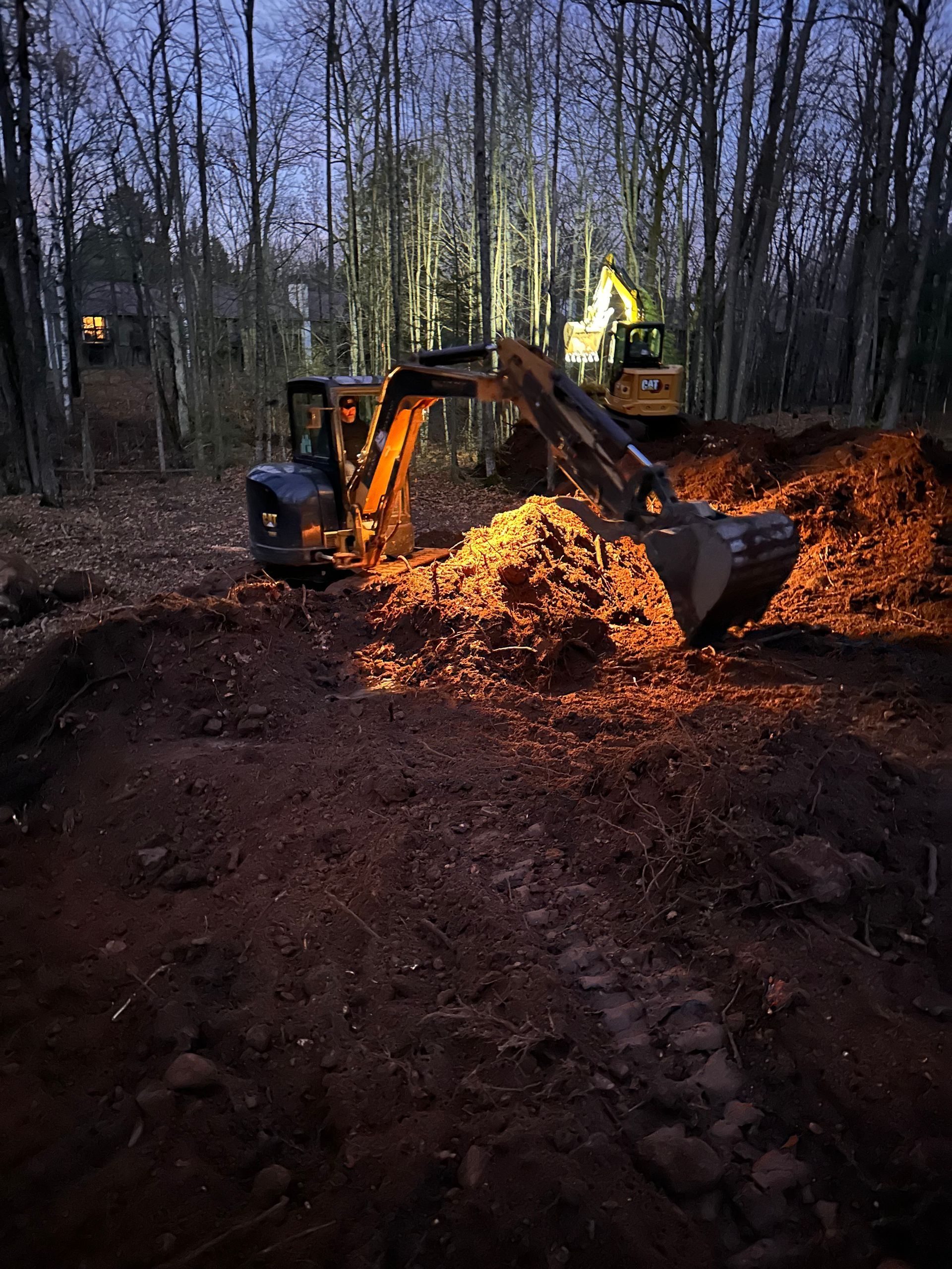 A yellow excavator at night works in a wooded area, scooping a large pile of dirt with its illuminated bucket. A yellow excavator at night works in a wooded area, scooping a large pile of dirt with its illuminated bucket.
