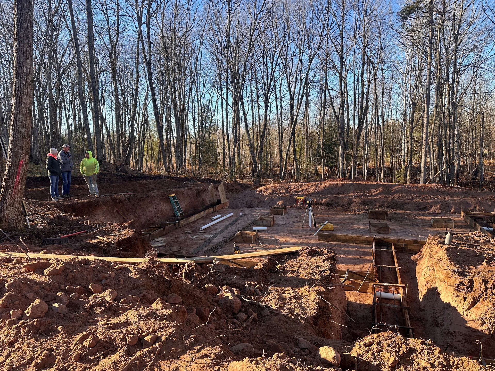 Three people stand on the edge of a dirt construction site with excavated foundation trenches in a wooded area.