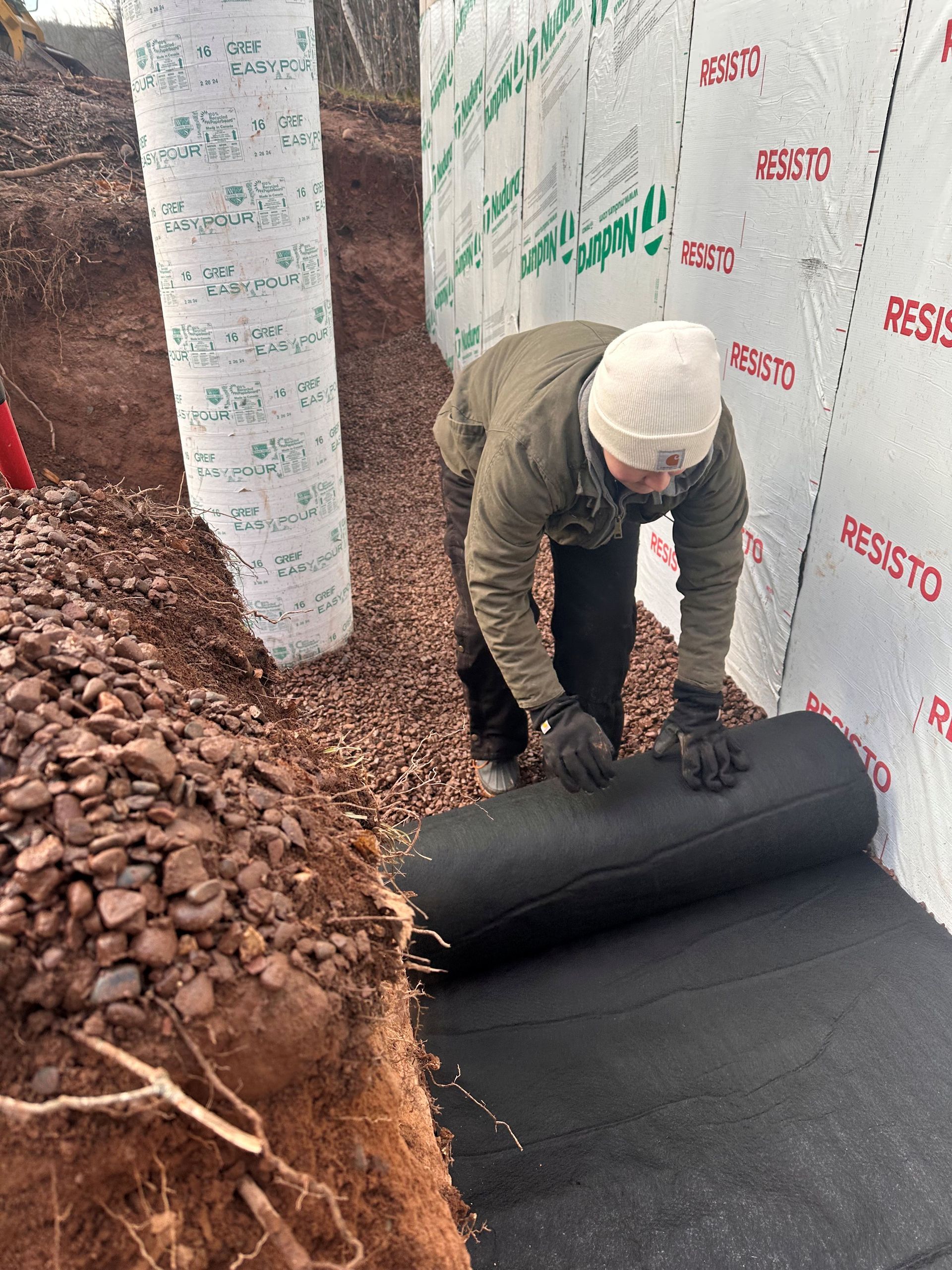 A person wearing a beanie and gloves unrolls black waterproofing membrane along the base of a foundation wall at a job site.