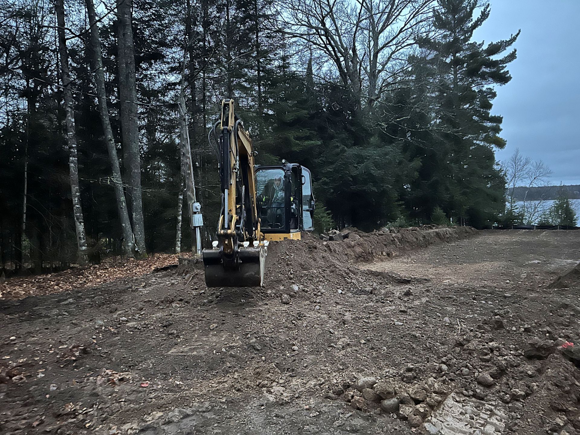 A yellow excavator sits in a cleared, gravelly lot near a treeline and a body of water under an overcast sky.