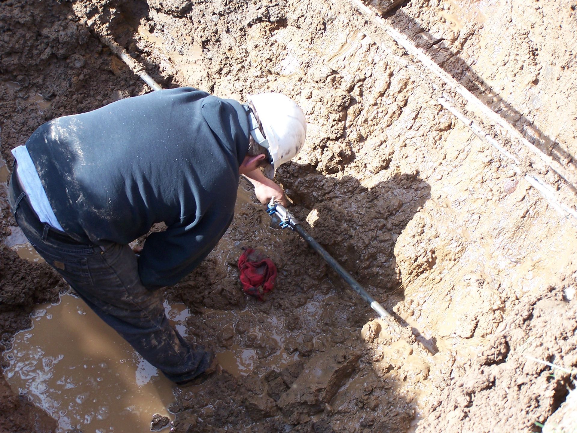 A person in a white hard hat and dark clothing works on a pipe in a muddy, excavated trench. A person in a white hard hat and dark clothing works on a pipe in a muddy, excavated trench.