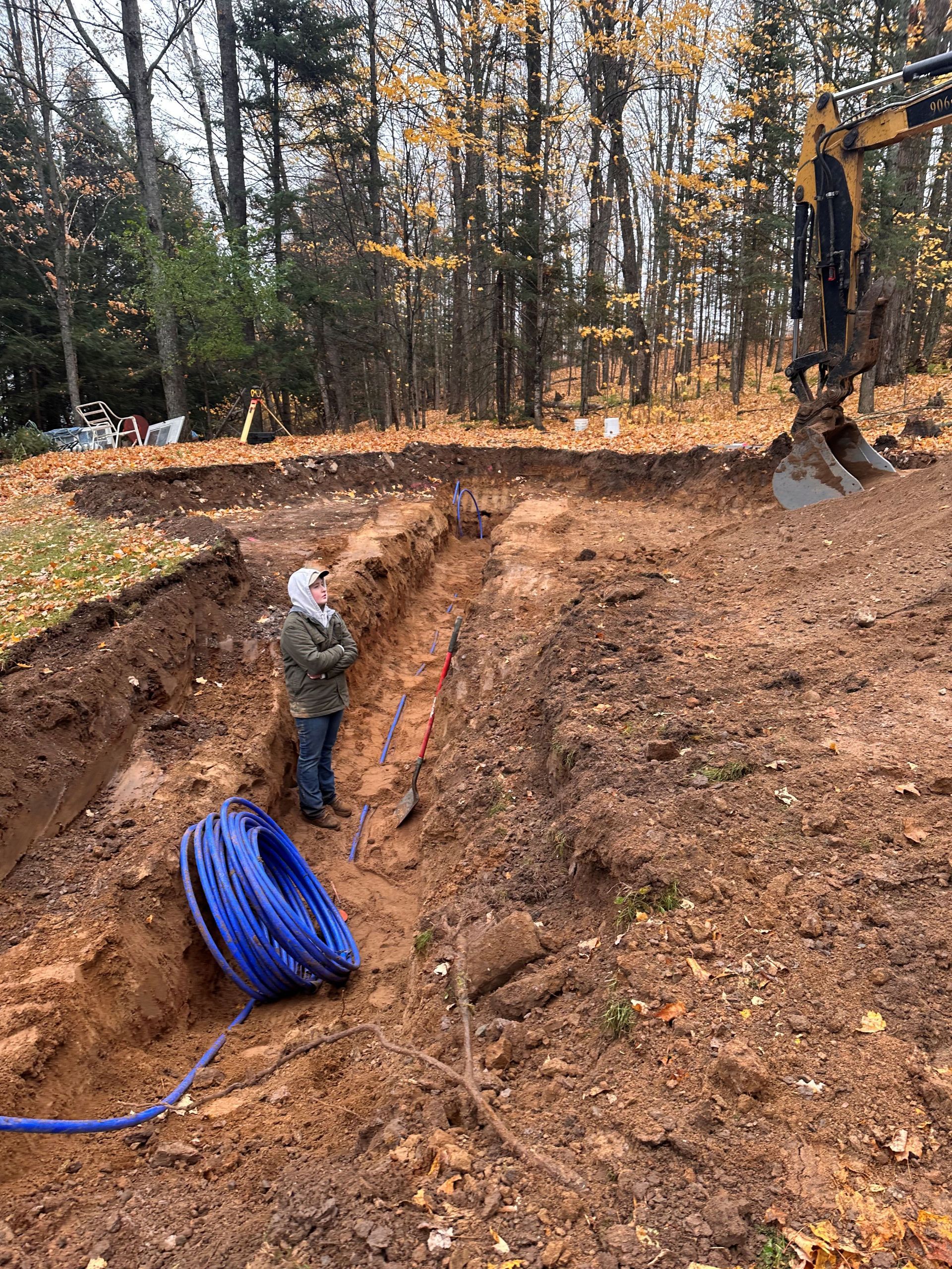 A person stands in a dirt trench in a wooded area near a blue plastic pipe coil and heavy machinery. A person stands in a dirt trench in a wooded area near a blue plastic pipe coil and heavy machinery.