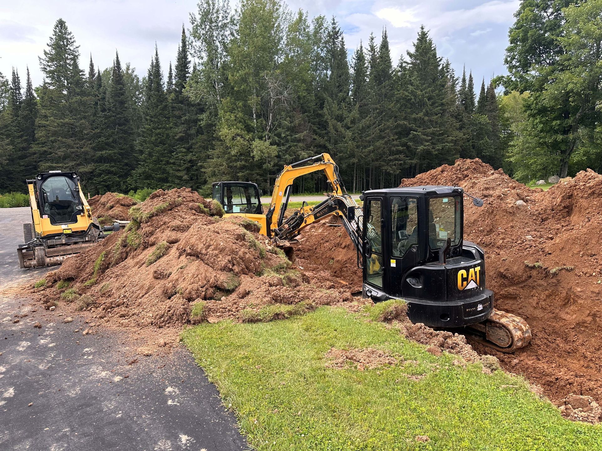 Two yellow and black Caterpillar excavators digging in piles of dirt near a wooded area and a paved driveway.