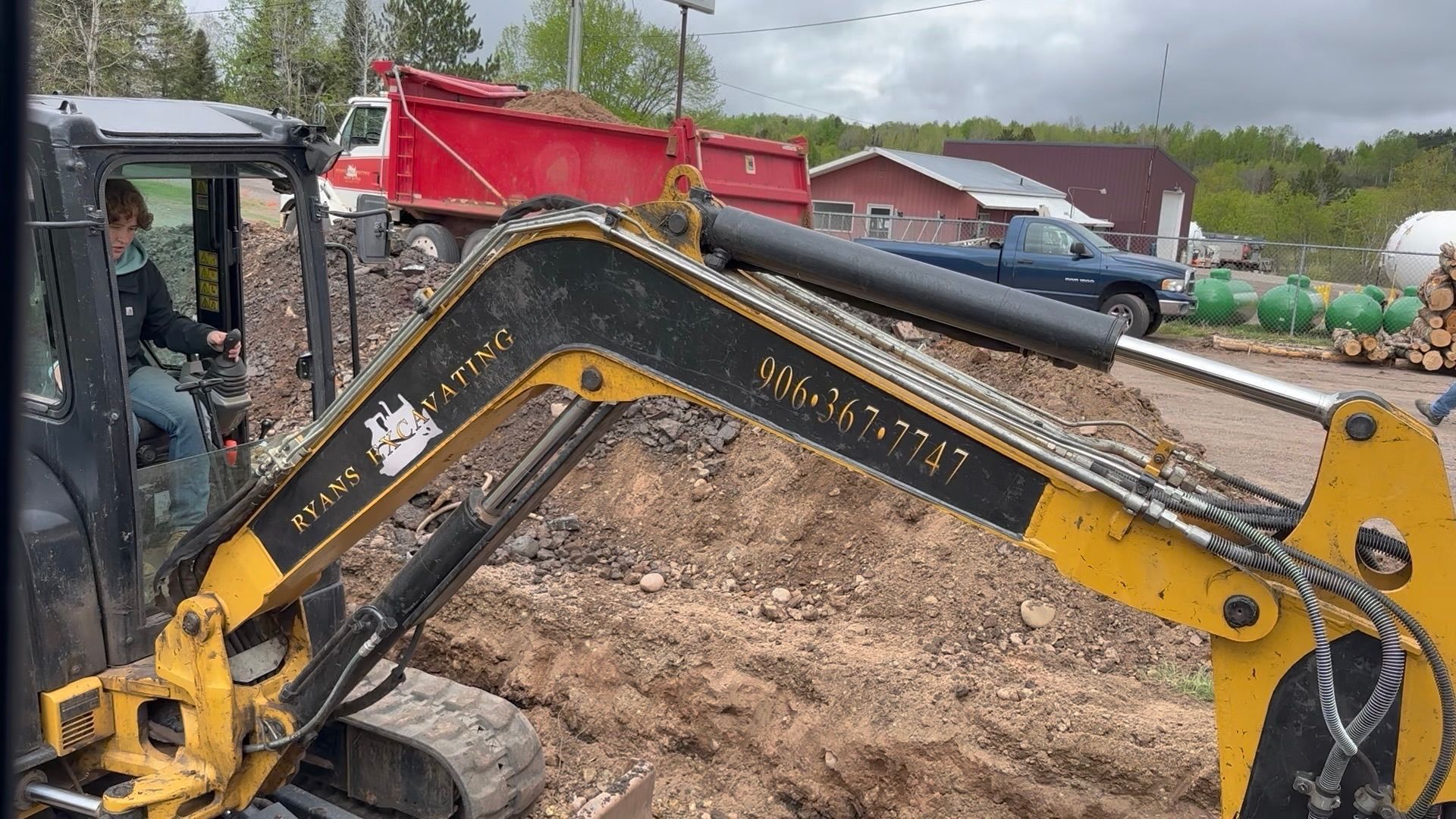 A person operates a yellow excavator digging a trench at an outdoor work site with a red dump truck in the background. A person operates a yellow excavator digging a trench at an outdoor work site with a red dump truck in the background.