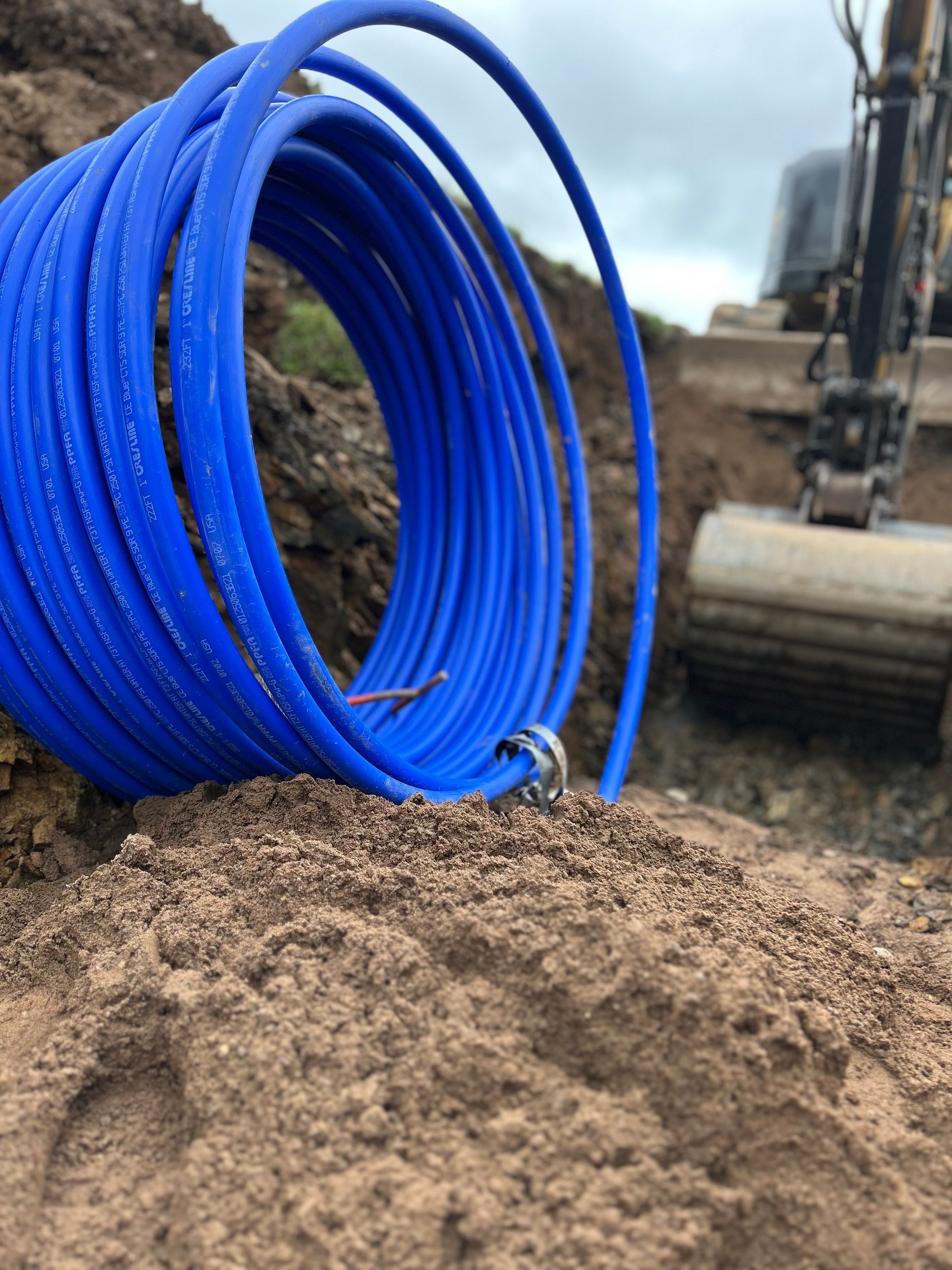 A coil of blue utility tubing rests on a pile of dirt in an outdoor construction site with an excavator in the background. A coil of blue utility tubing rests on a pile of dirt in an outdoor construction site with an excavator in the background.