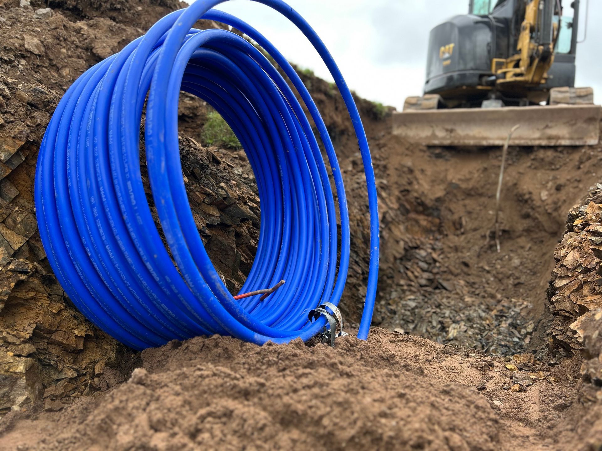 A large coil of blue irrigation pipe sits on the dirt edge of an excavation trench with construction machinery nearby.