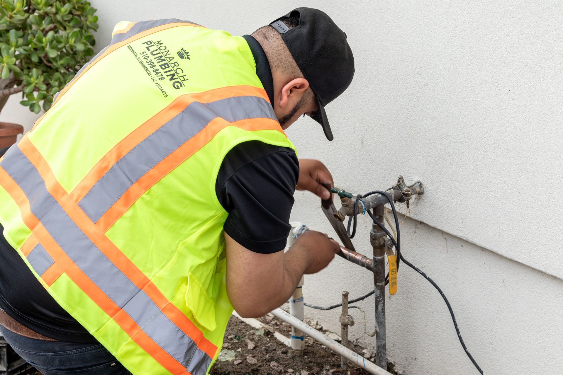 Worker in a neon safety vest repairs outdoor utility piping beside a building.
