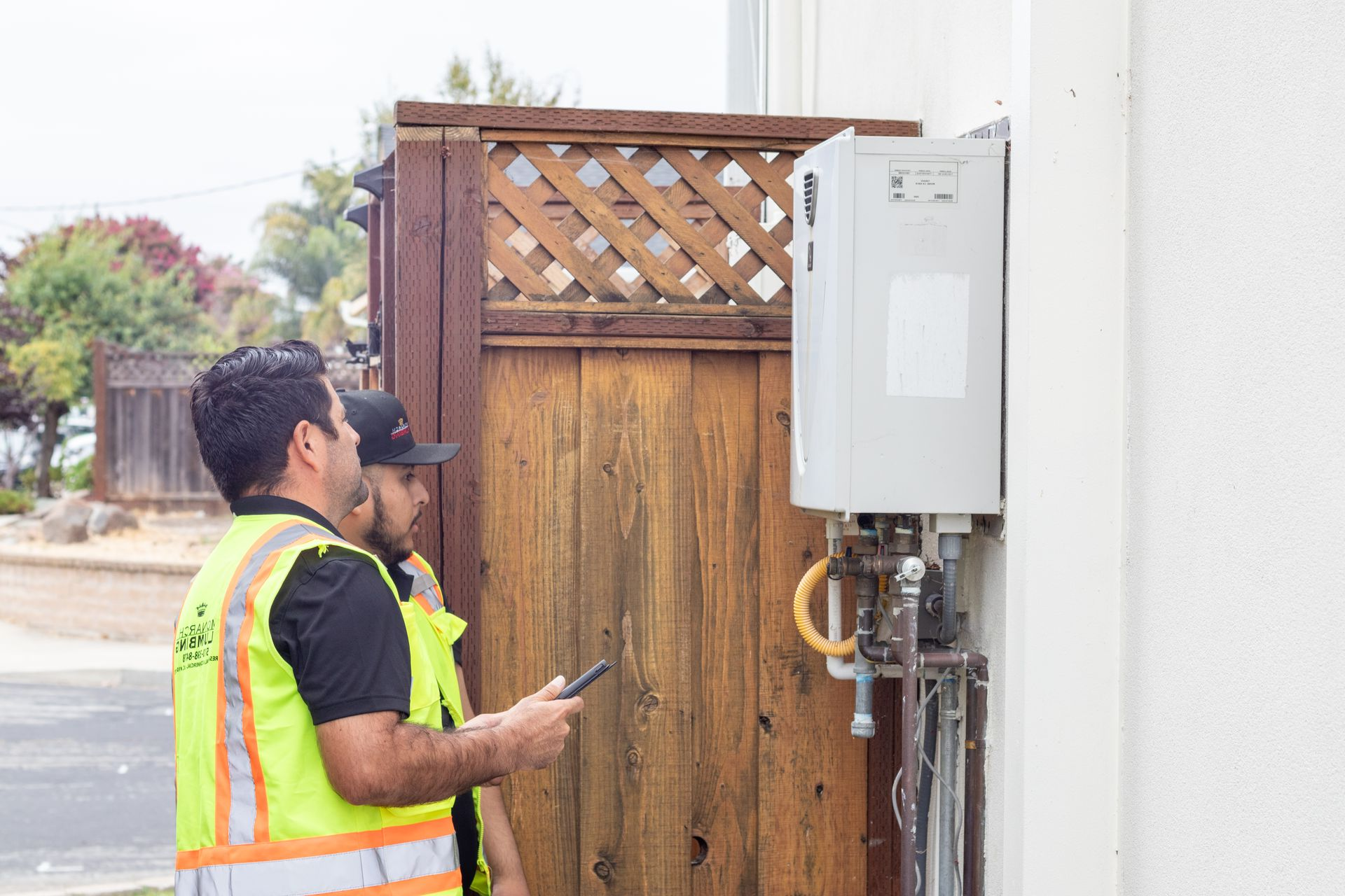 Two workers inspecting a a tankless water heater outside near a wooden fence