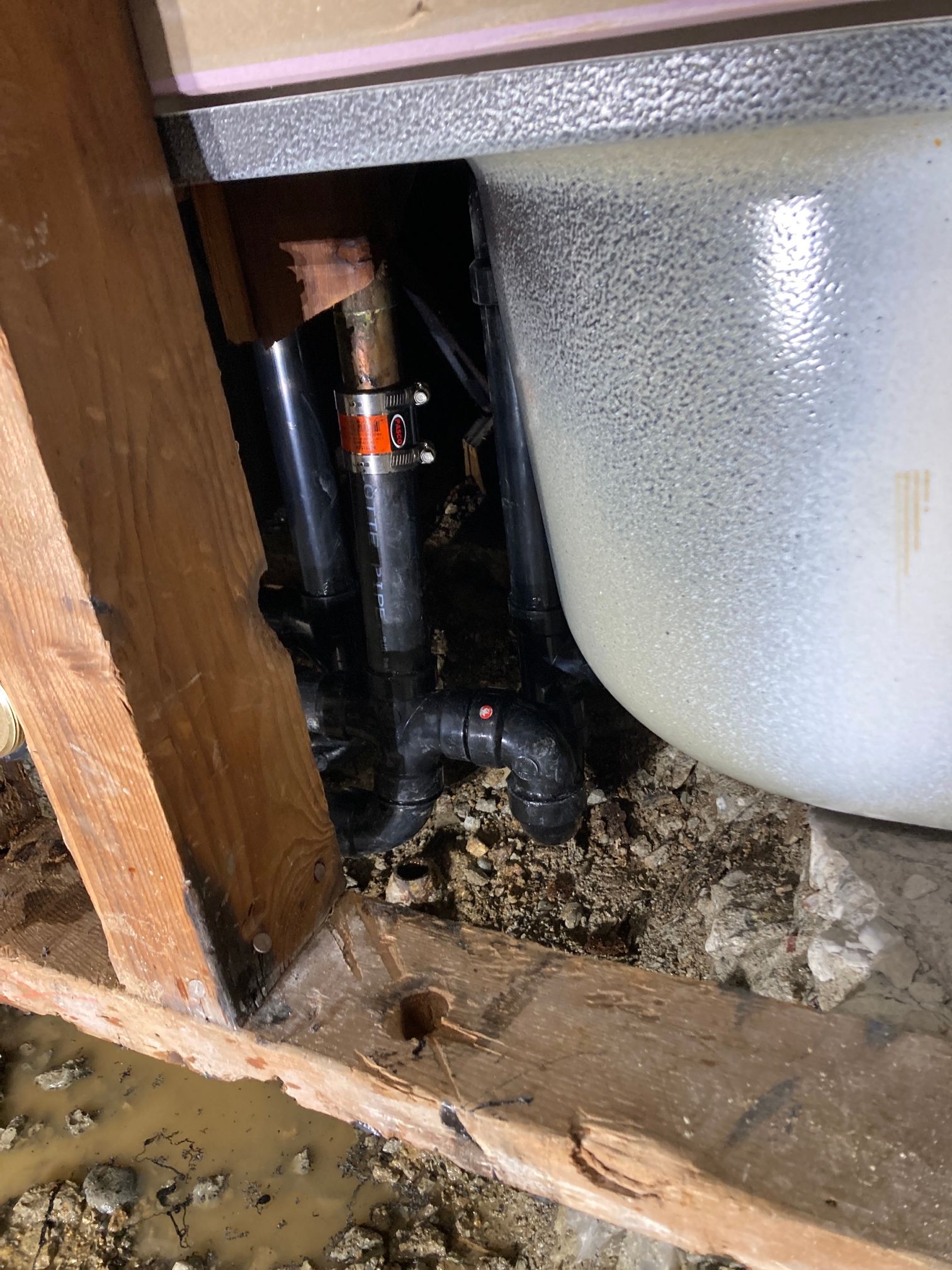 Close-up of plumbing under a sink, with a black pipe, copper fitting, and white basin in a wooden cabinet.
