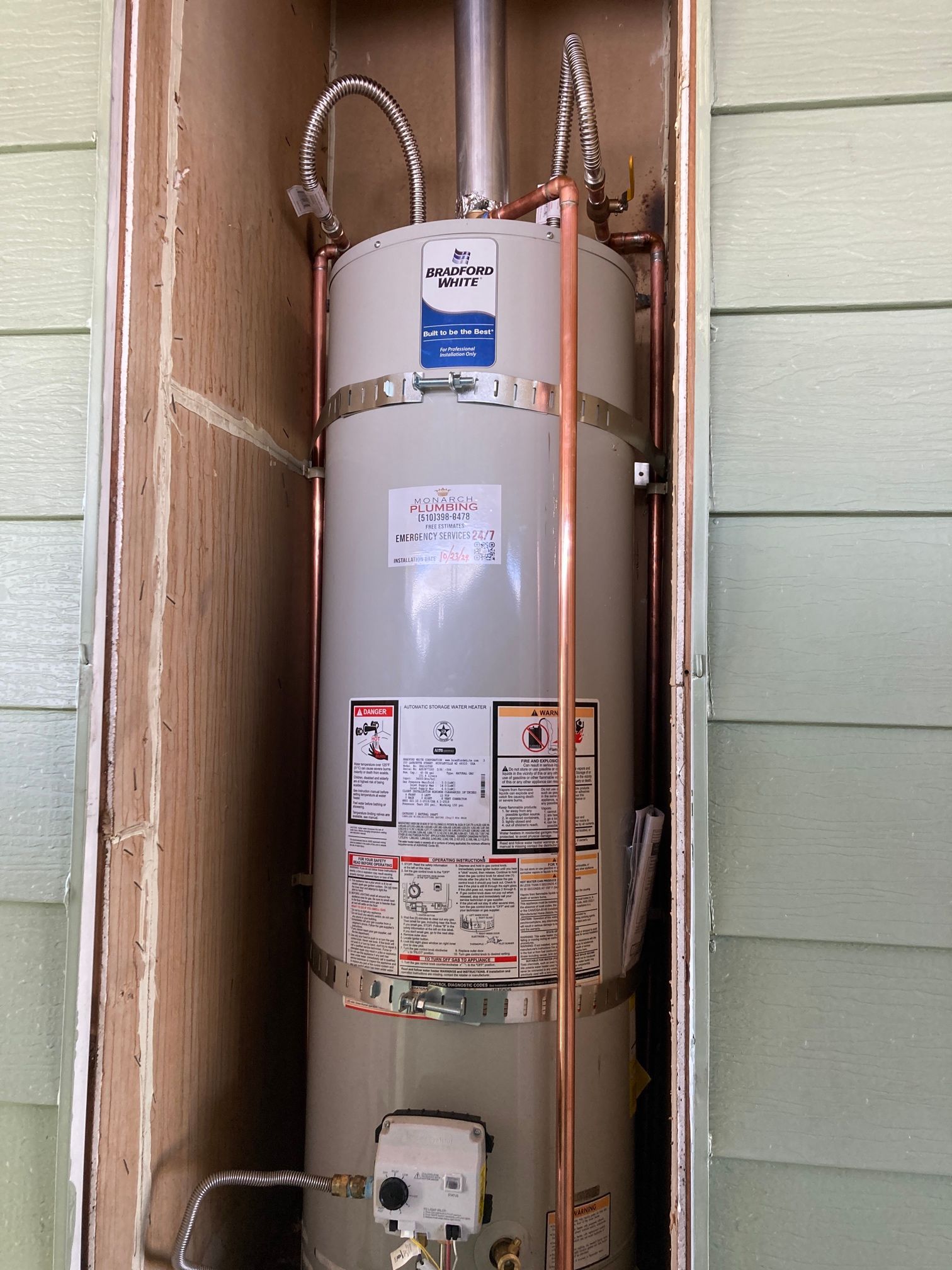 Tall water heater in an outdoor alcove with copper pipes.