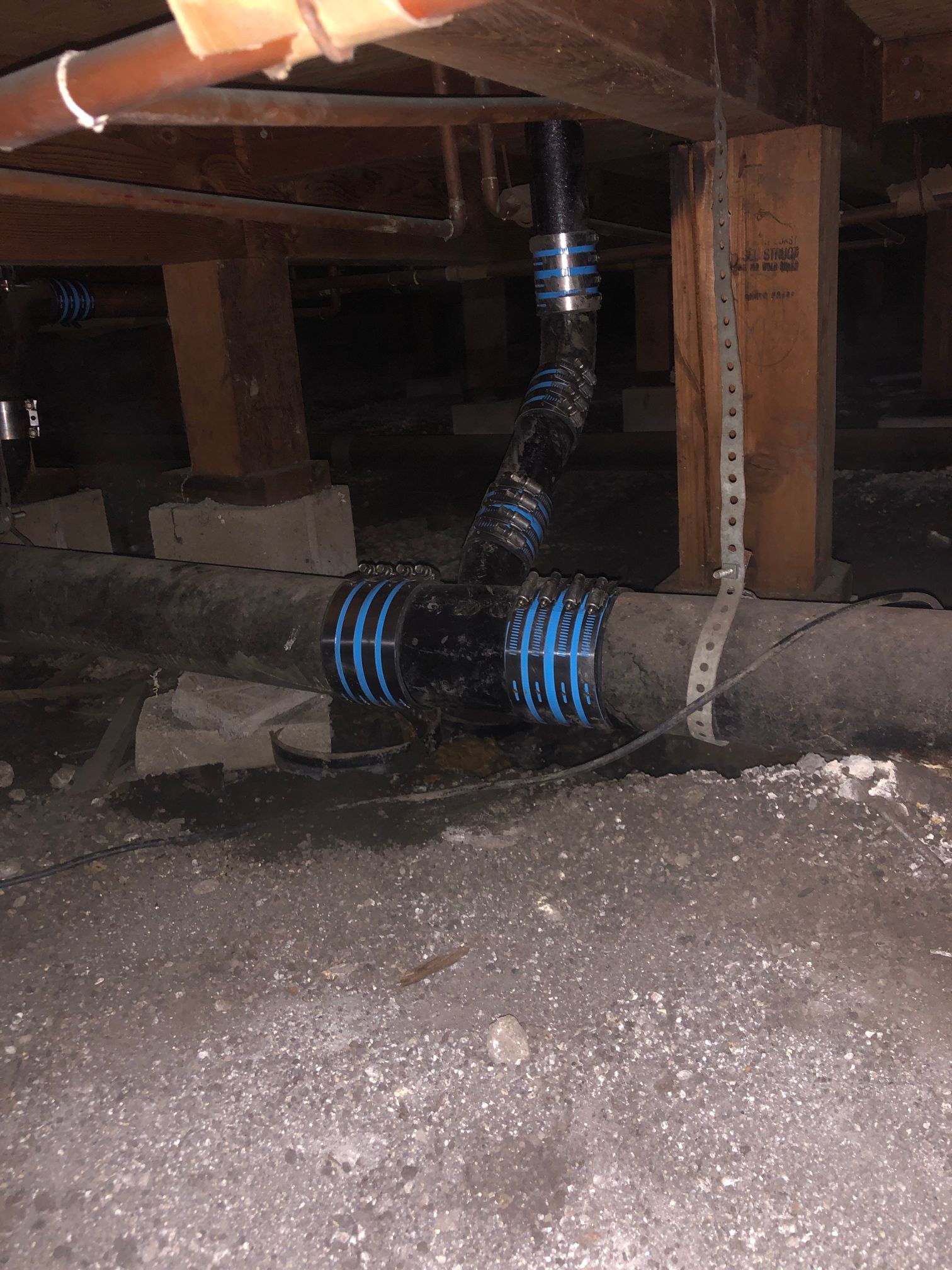 Crawlspace with a black corrugated pipe wrapped in blue rings and insulation beneath wooden joists