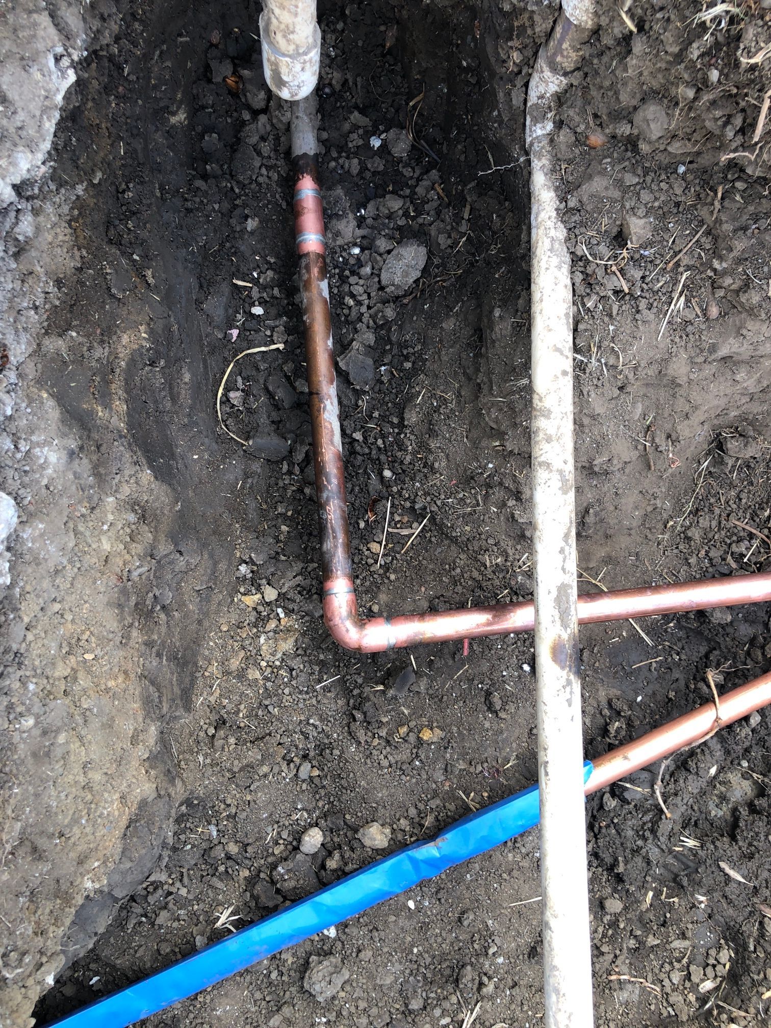 Trenched ground with exposed copper pipes and a blue pipe crossing the dirt.