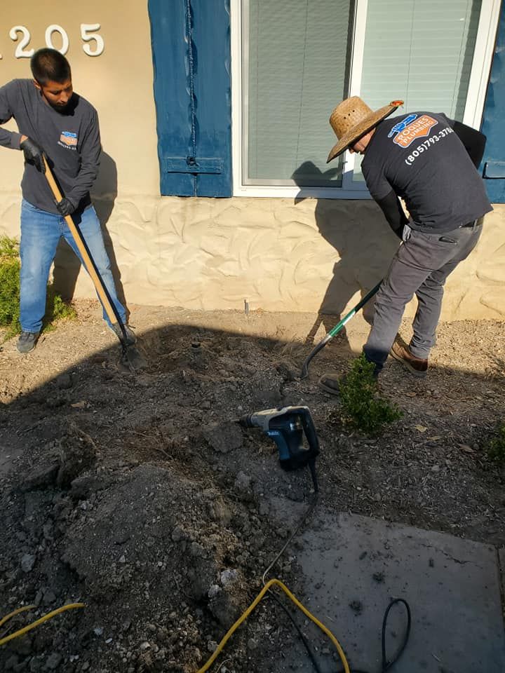 Two men are digging in the dirt in front of a house.