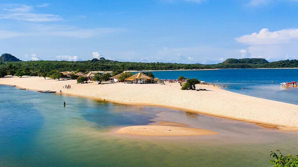 Sandy beach peninsula with calm, blue water, tropical trees, and buildings under a bright sky.