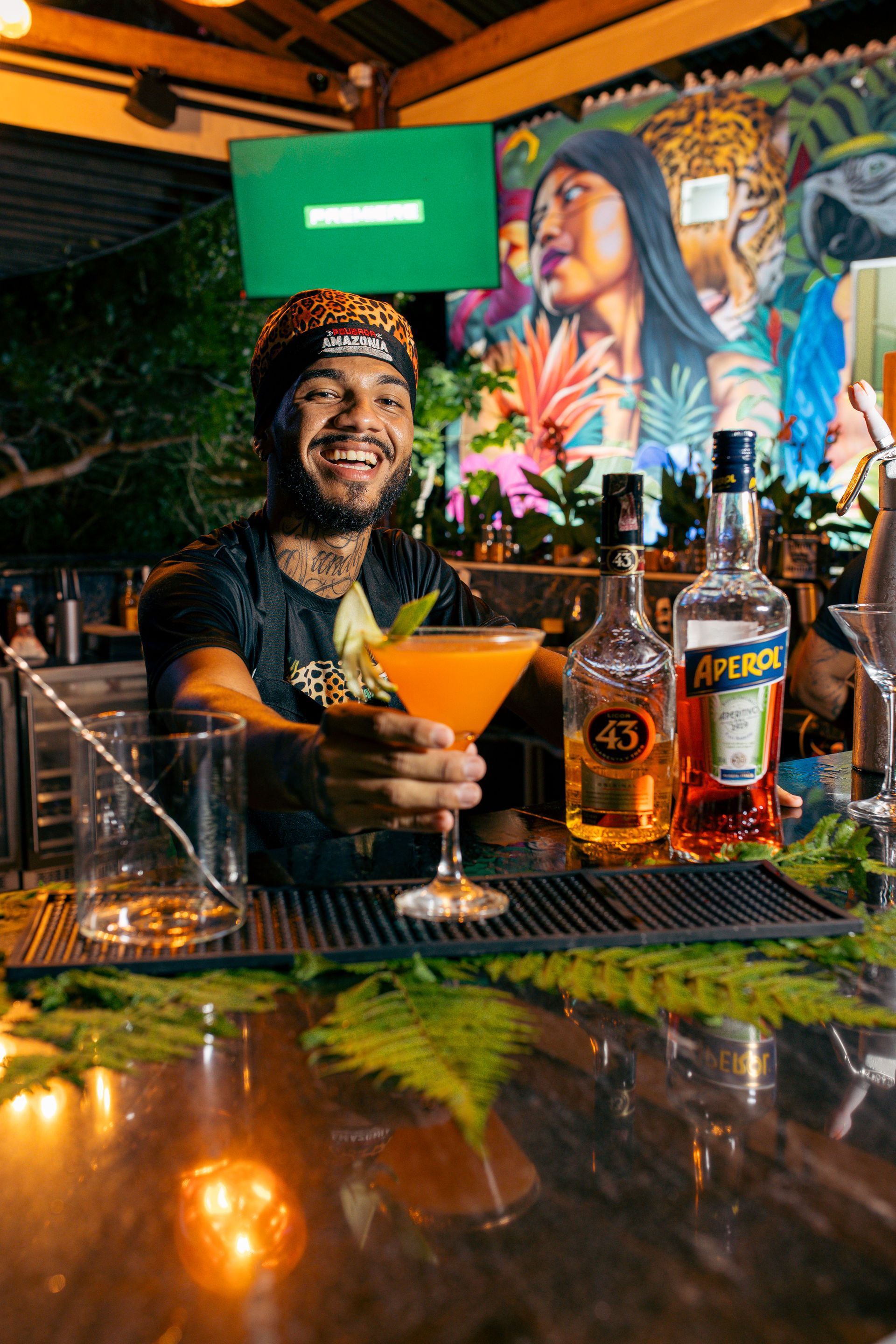 Smiling bartender holds a cocktail; bar with liquor bottles. Colorful mural in the background.