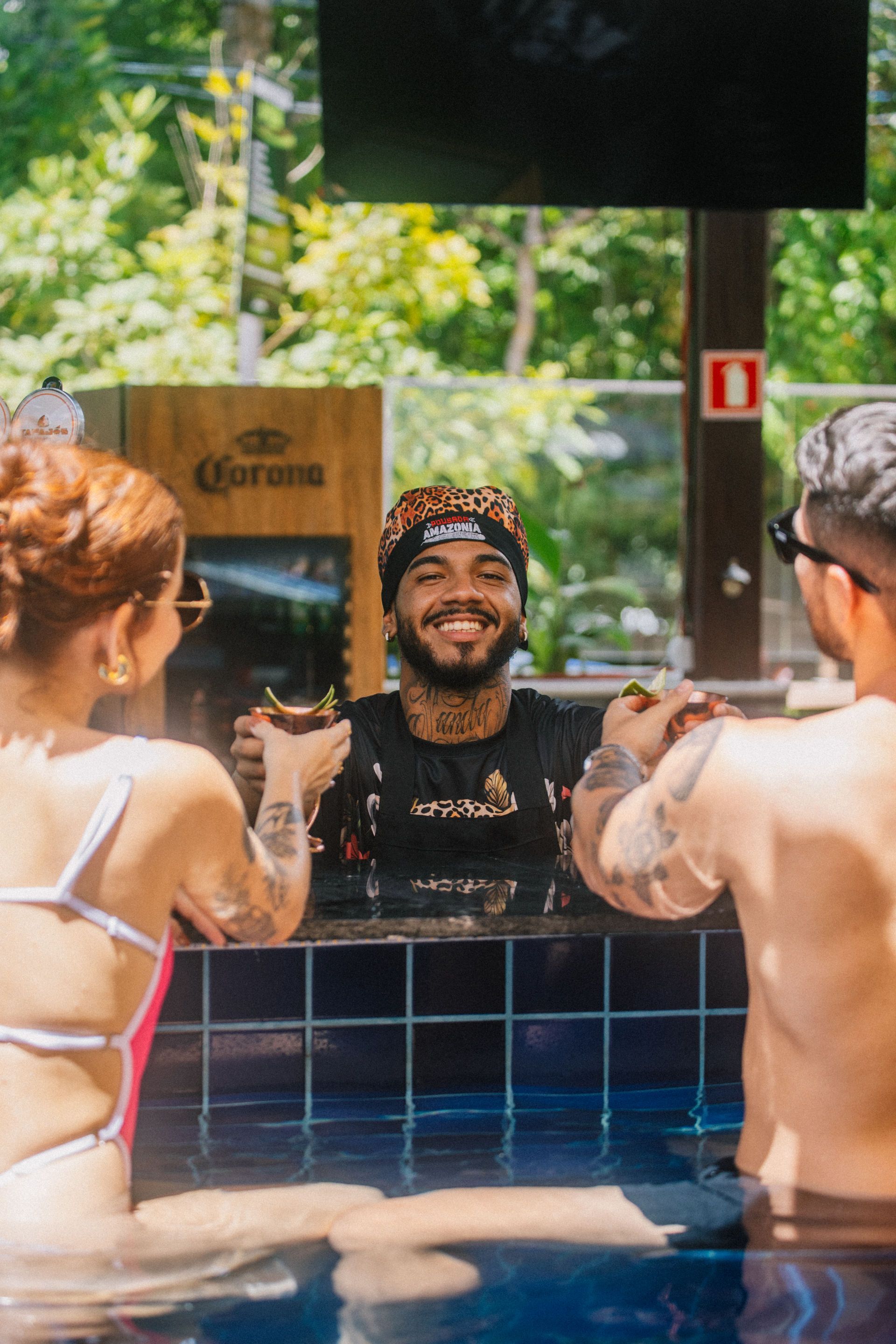 Bartender smiles, serving drinks to two people in a pool. Sunny outdoor setting.
