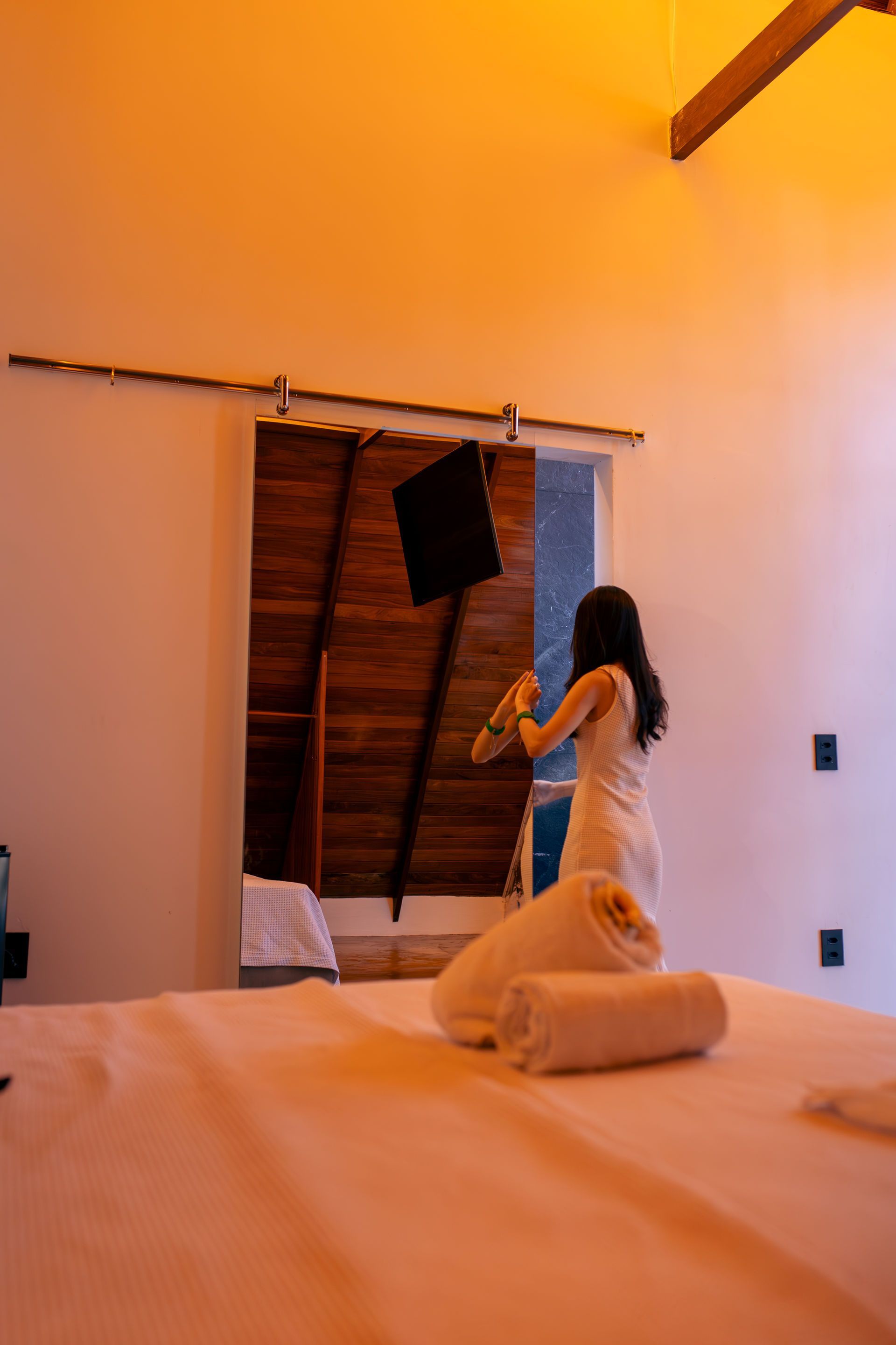 Woman in white dress by a mirror door, indoors. White bed with rolled towels. Warm lighting.