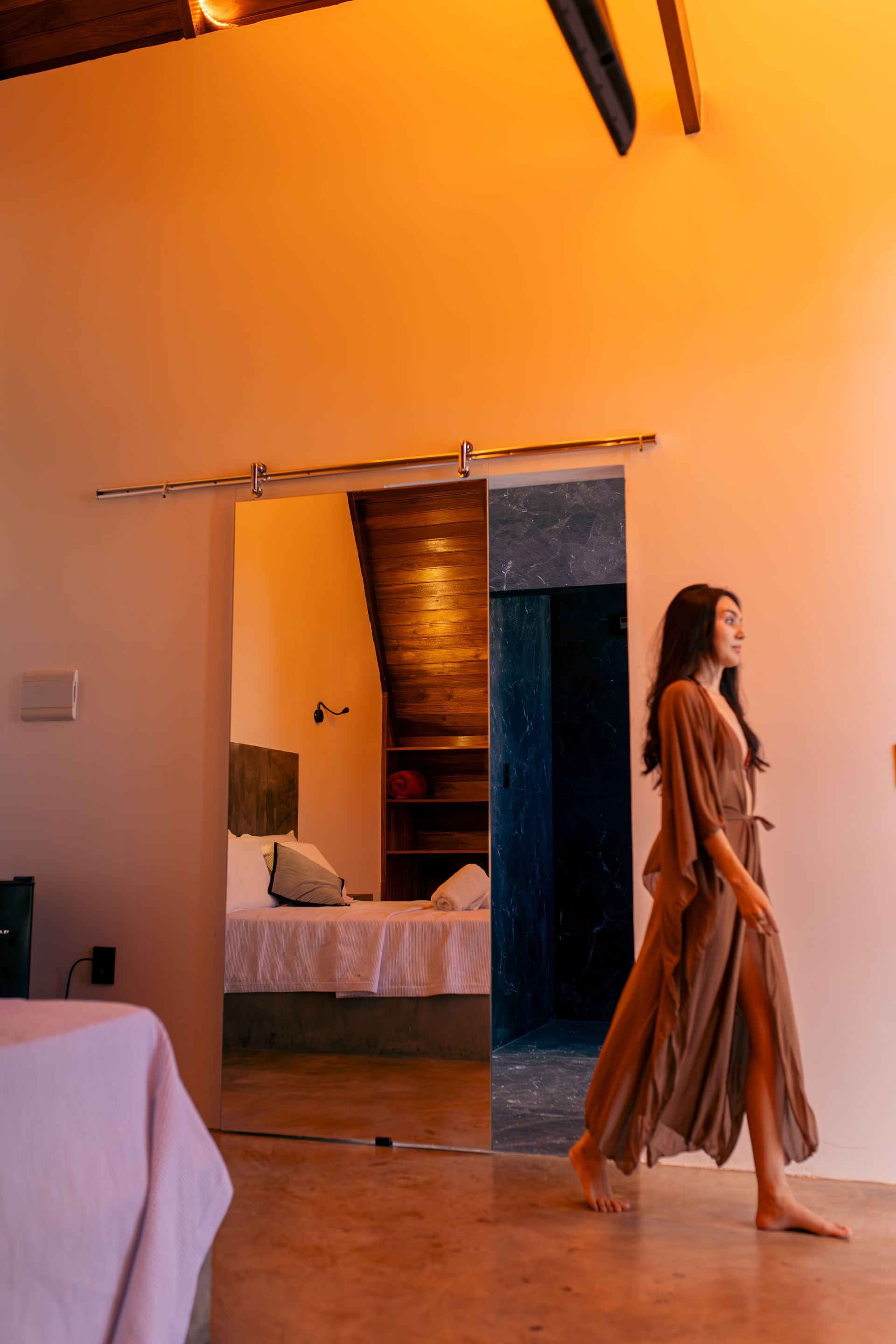 Woman in brown robe walks through a mirrored doorway to bedroom.
