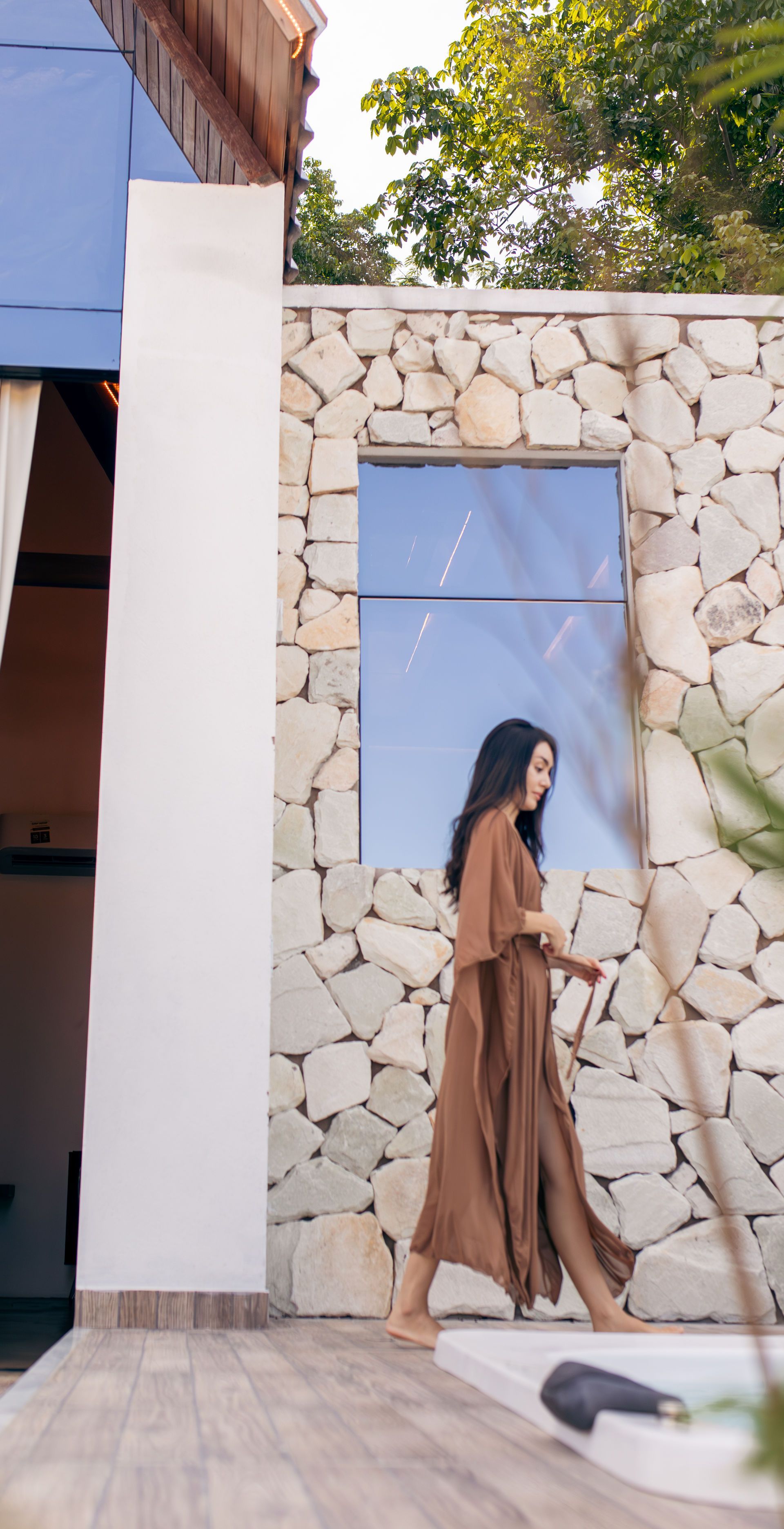 Woman in brown dress walking barefoot near stone wall and window.