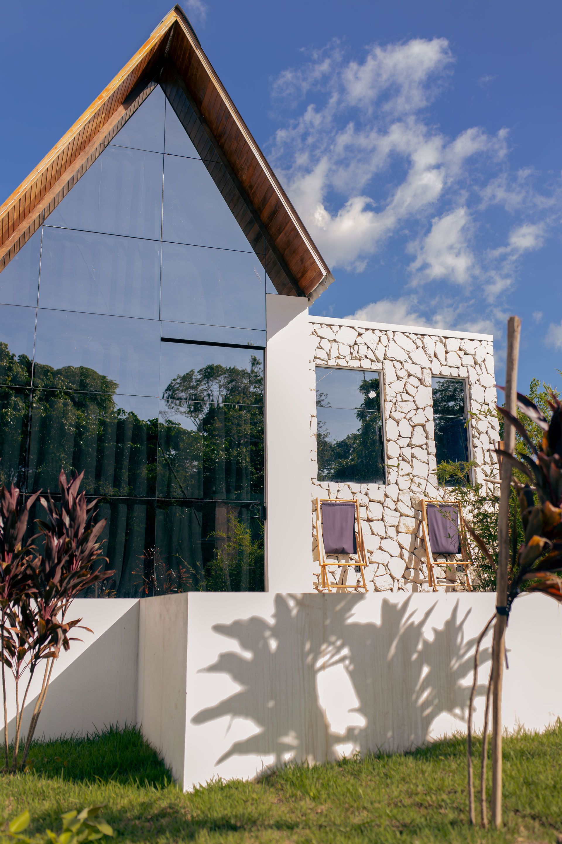 Modern white house with glass facade and wood trim, reflecting trees. Two chairs on balcony.