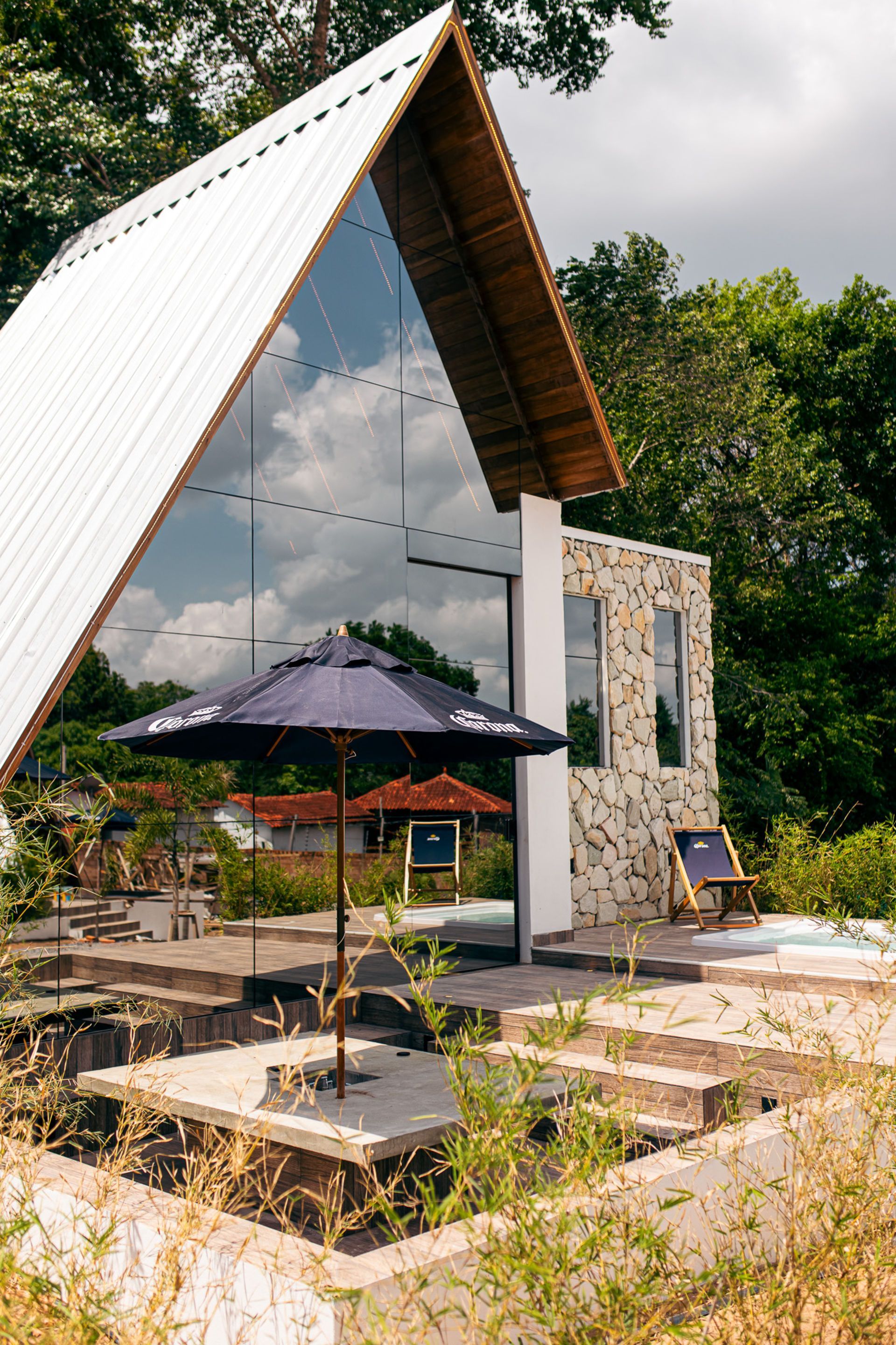 Modern cabin with glass facade, umbrella, and stone wall amidst greenery.