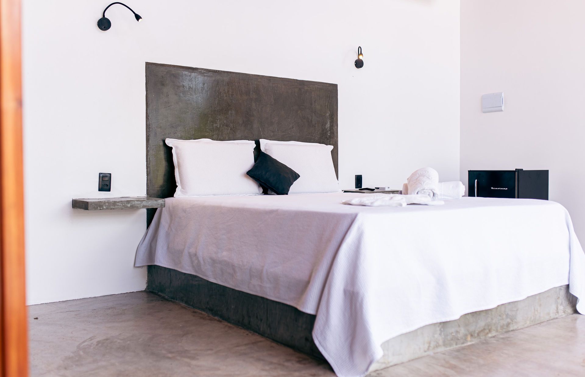 Bedroom with bed, concrete headboard, white bedding, black pillow, and side table.