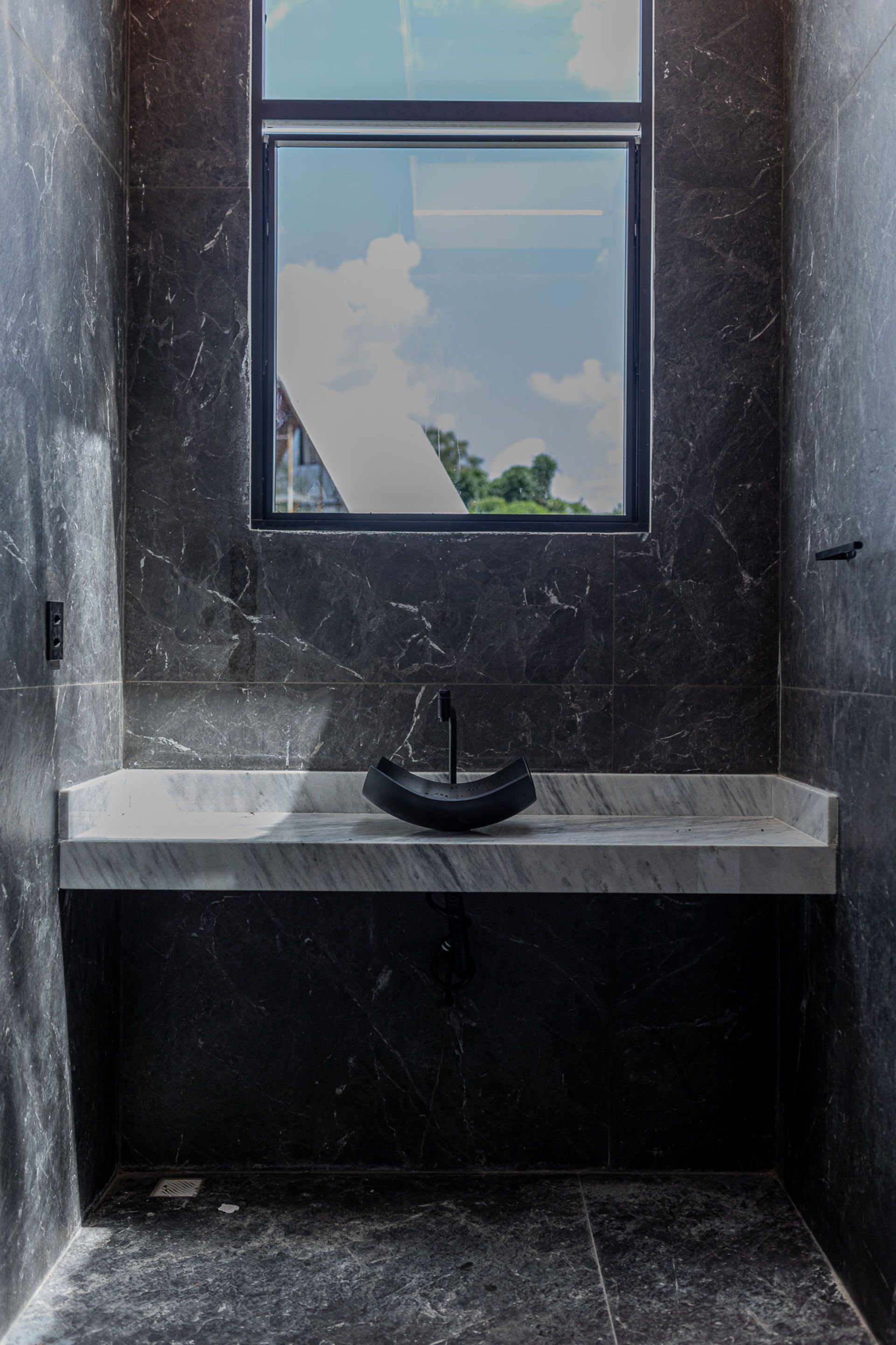 Bathroom with black marble walls, marble countertop, dark vessel sink, and window with sky view.