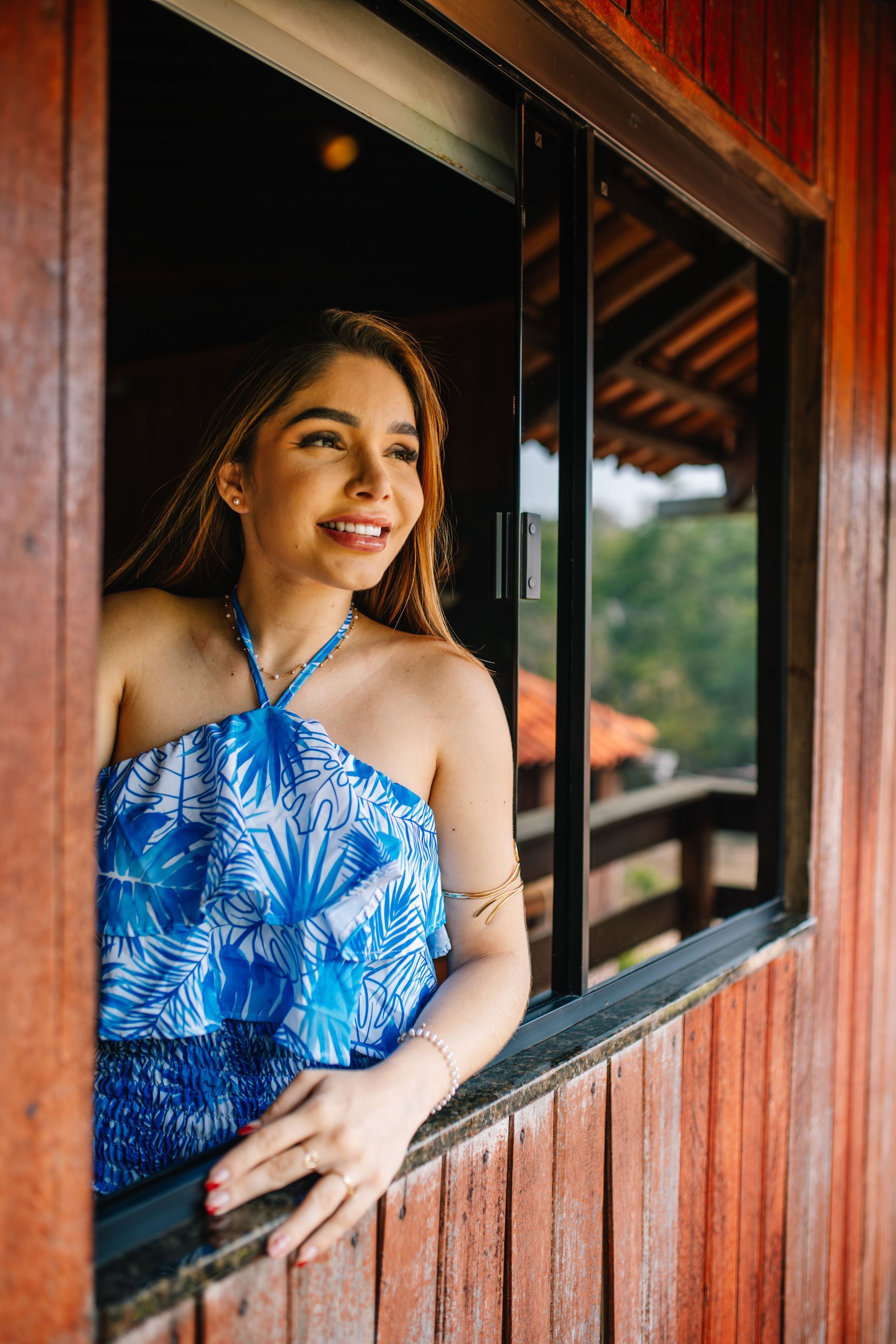 Woman smiling in a window, wearing blue floral top, looking outside; wooden cabin exterior.
