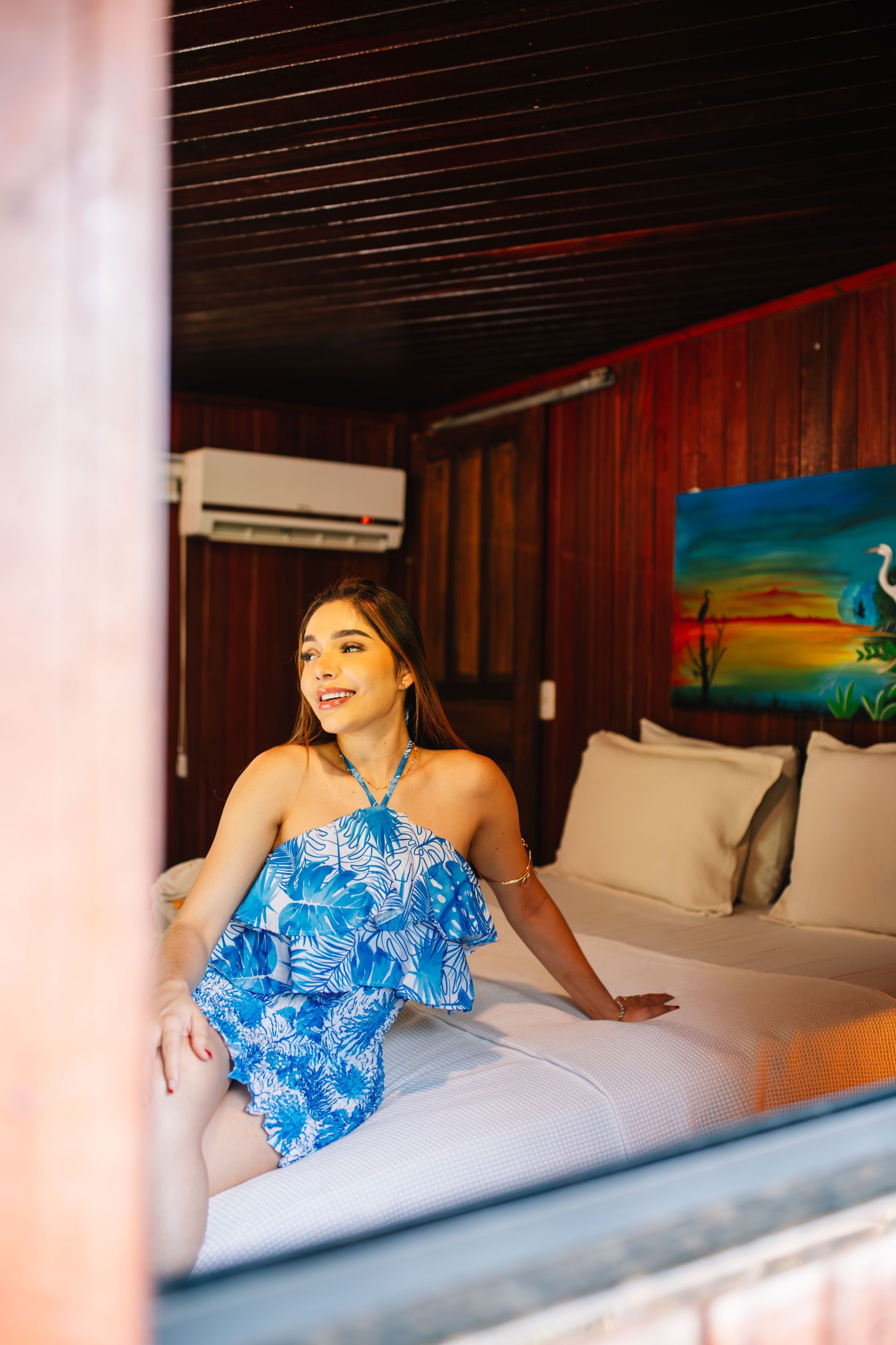 Woman in blue dress smiles on a bed in a wooden room, looking out a window.