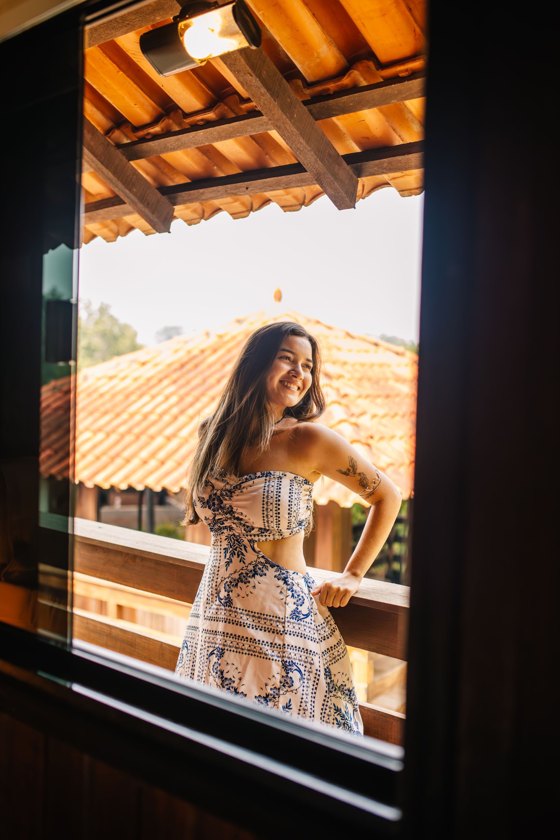 Woman in floral outfit smiles on a wooden balcony overlooking tiled rooftops.