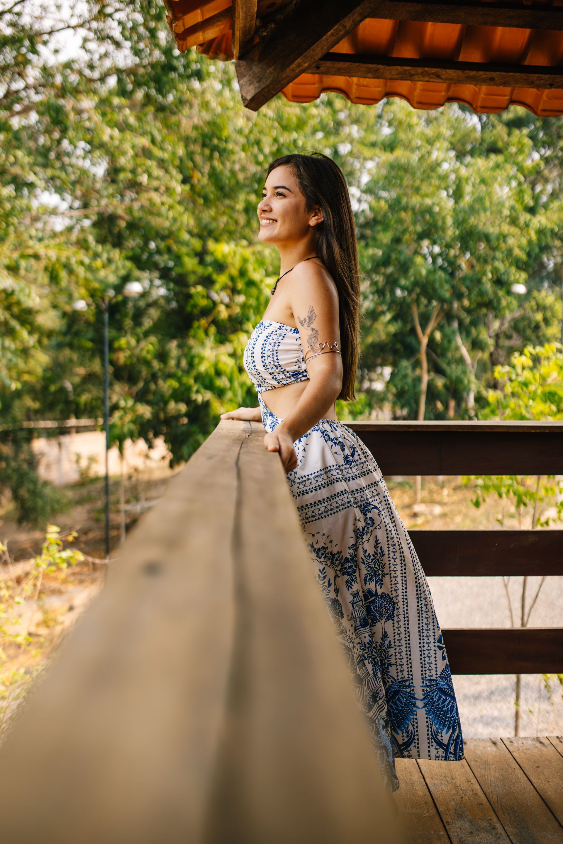 Woman in a blue and white dress, leaning on a wooden railing, looking out at a green landscape.
