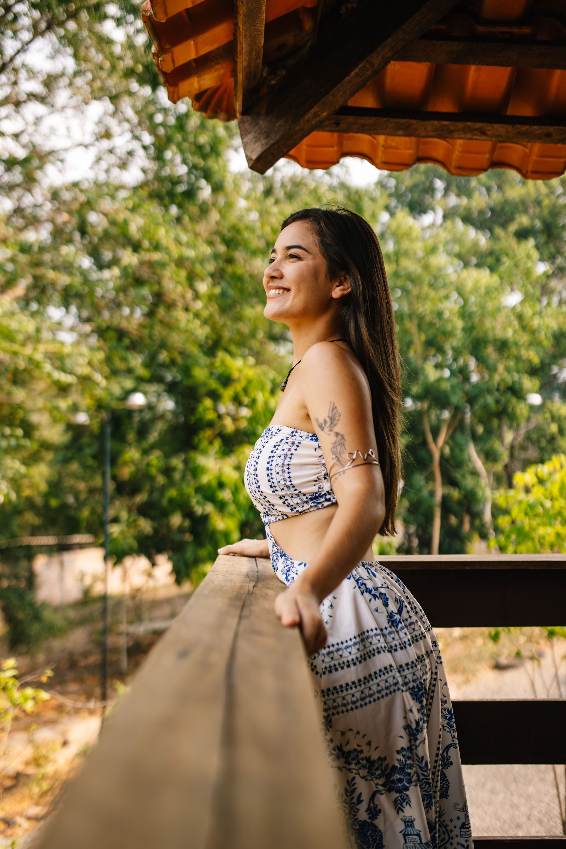 Woman in a white and blue patterned outfit smiles, leaning on a wooden railing, with trees in the background.