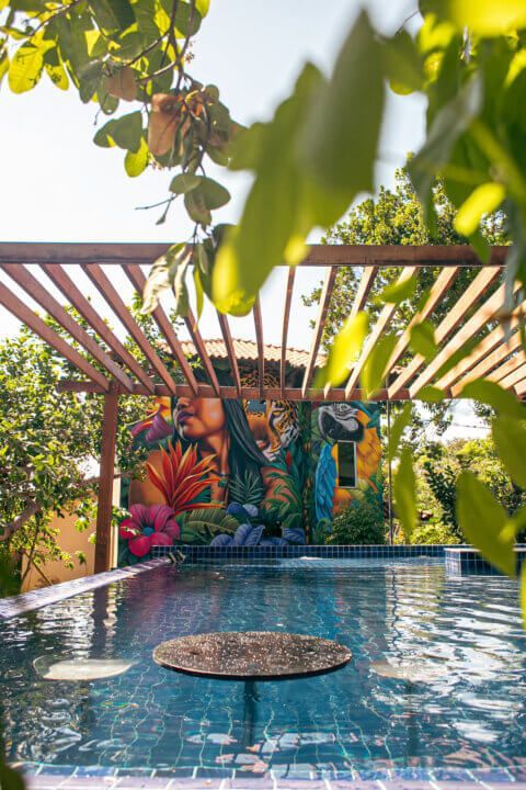 Swimming pool with a mural of a woman and flowers; wooden pergola overhead, framed by greenery.