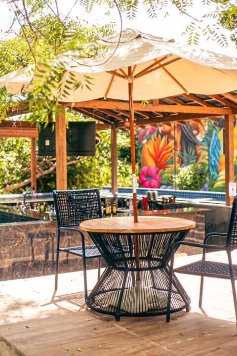 Outdoor dining table with umbrella, black chairs, tropical mural background.