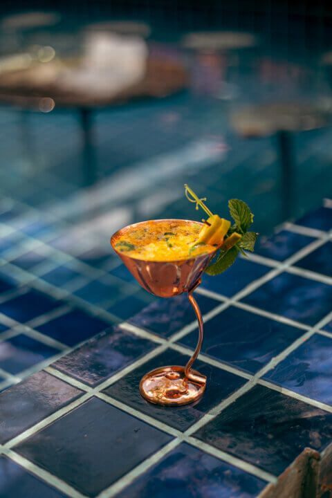 Golden cocktail in copper glass with mint garnish on blue tiled surface near a pool.