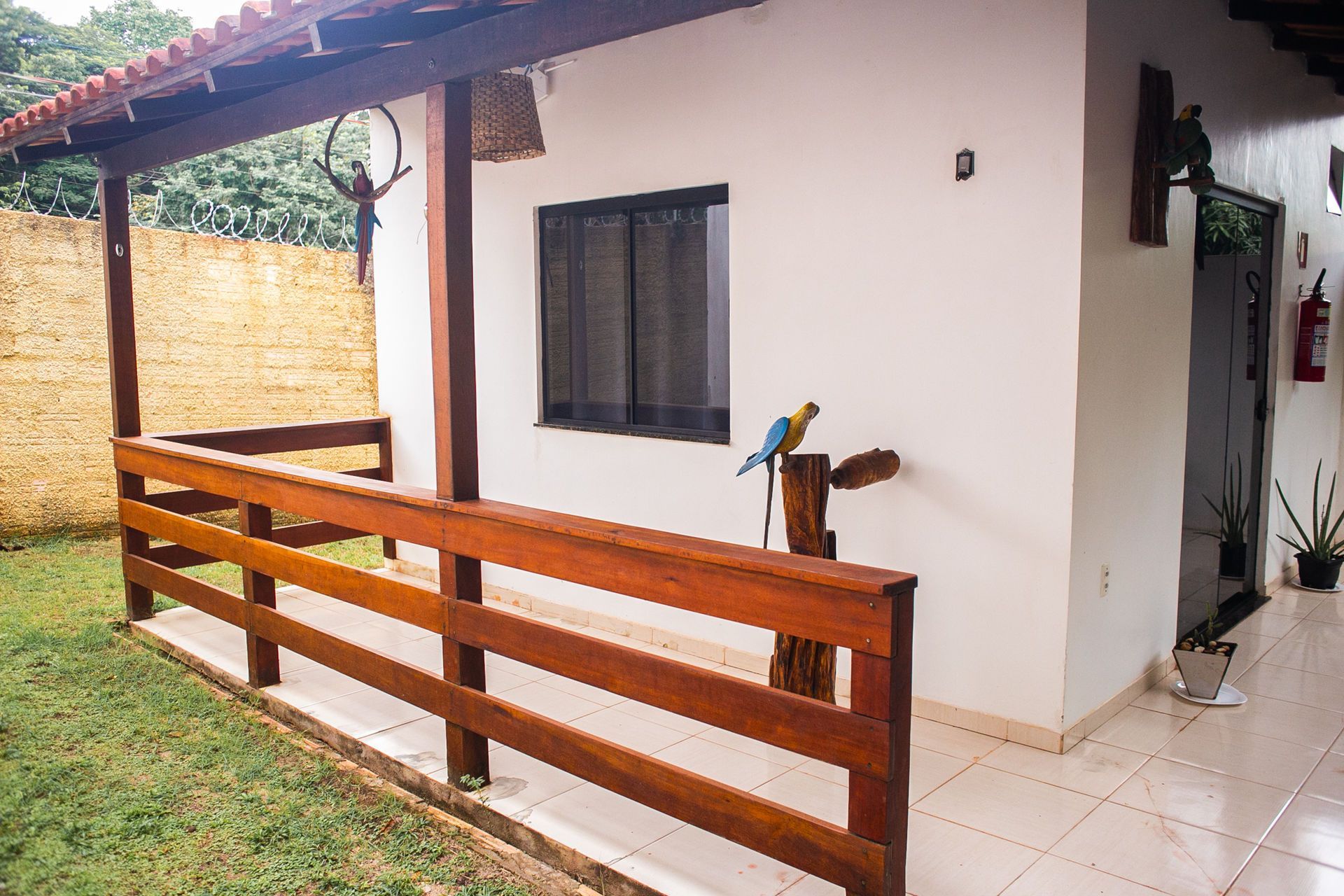 White cottage with a wooden porch and railing; lawn and trees in the background; a macaw statue is perched on a support post.